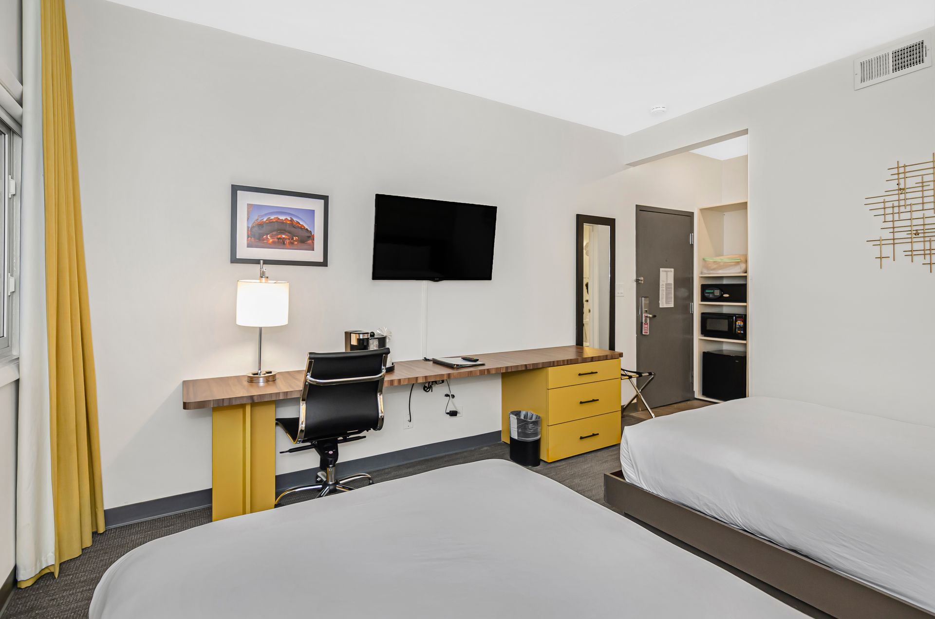 Hotel room with two beds, a desk with a black office chair, a wall-mounted TV, and a microwave on a shelving unit.