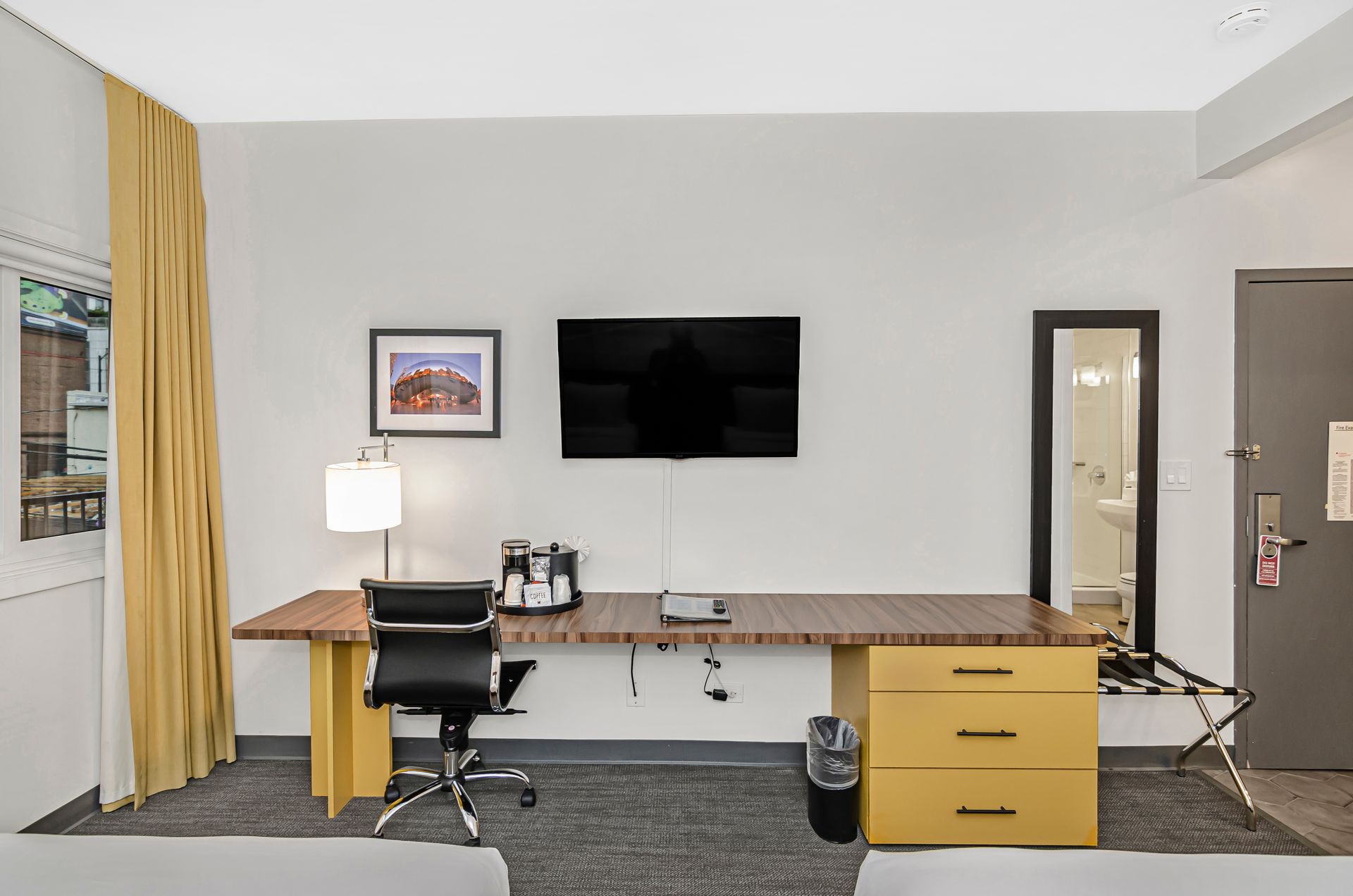 A hotel room desk area with a wall-mounted TV, office chair, desk lamp, coffee maker, and three-drawer cabinet.