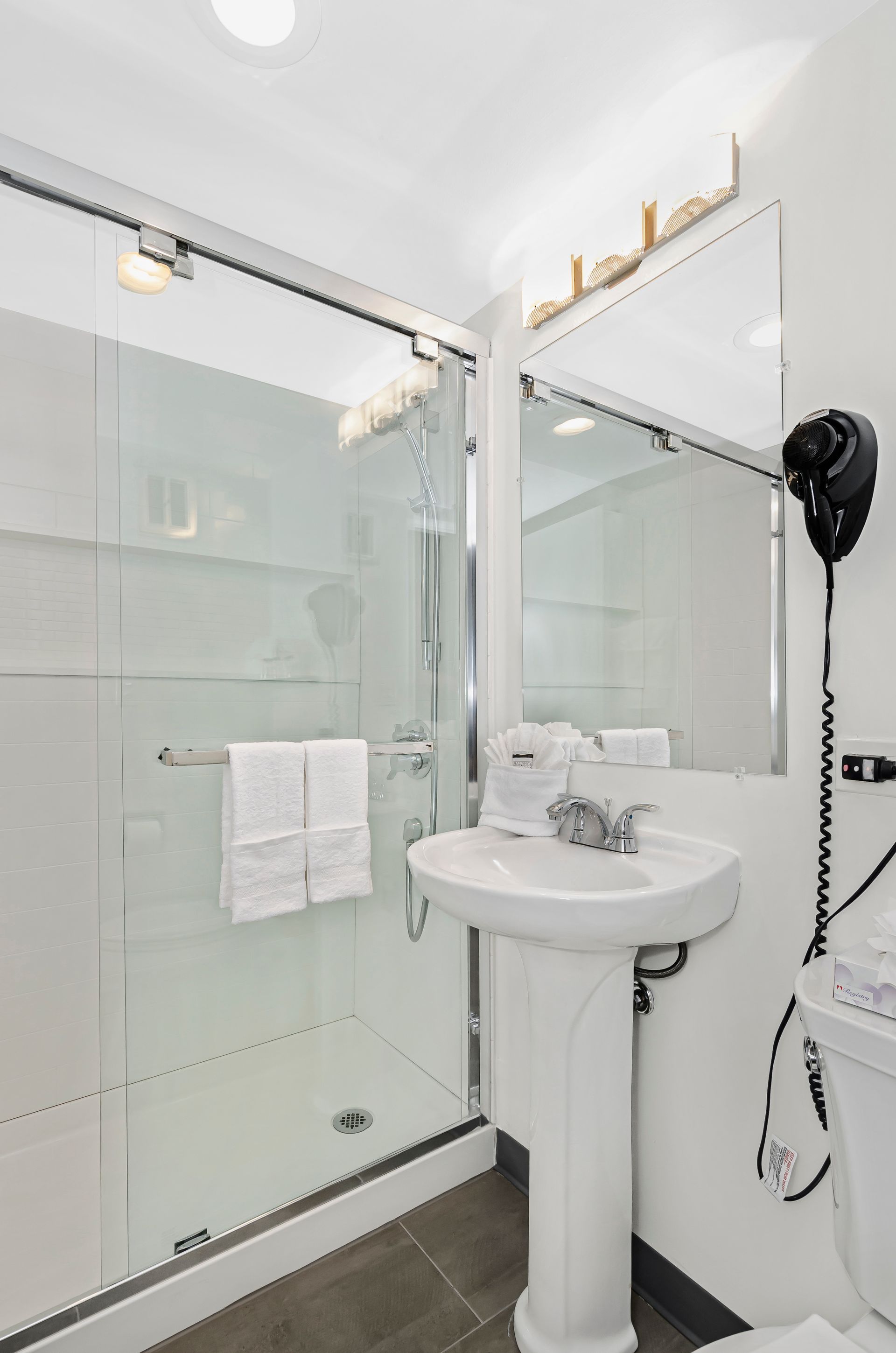 A compact bathroom featuring a glass-enclosed shower, a white pedestal sink, a rectangular mirror, and a wall-mounted dryer.