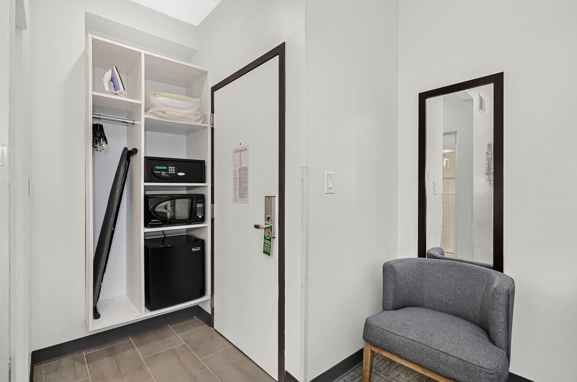 A hotel closet area with white shelves holding an iron, towels, and a mini-fridge, next to a chair and a wall mirror.