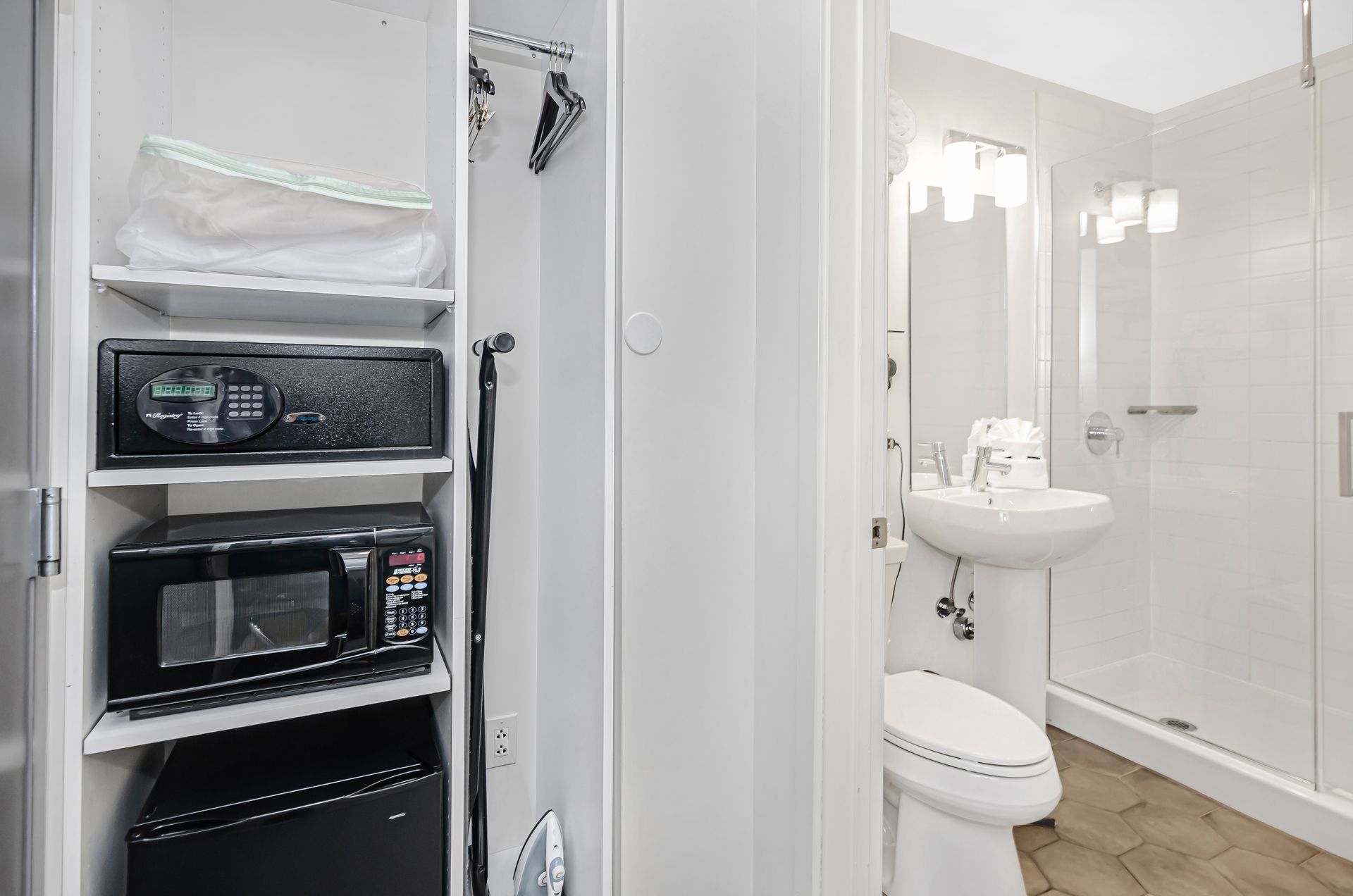 A hotel room alcove with a mini-fridge, microwave, and safe, next to a white bathroom featuring a sink, toilet, and shower.