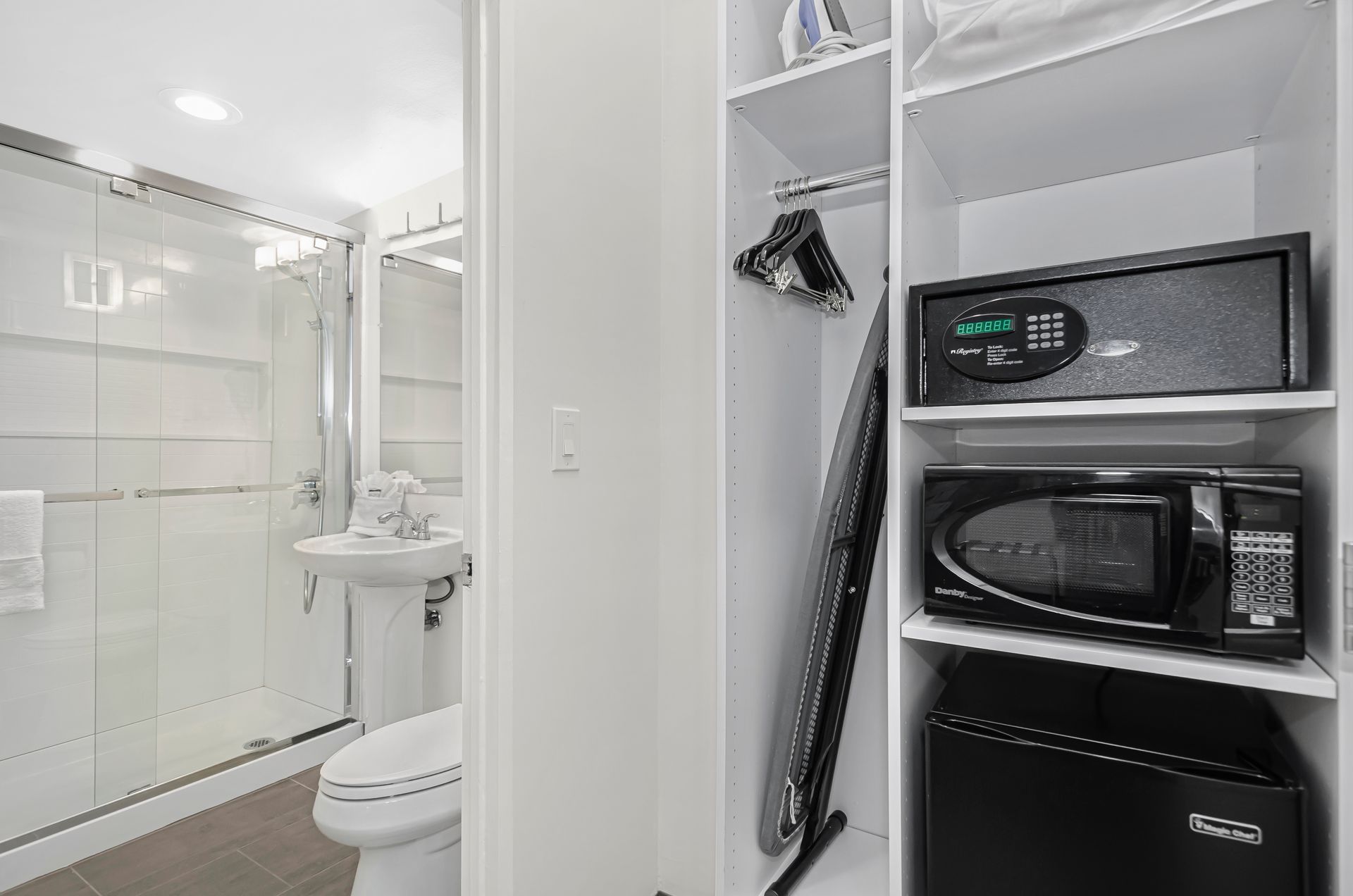A white-walled hotel closet containing a safe, microwave, and mini-fridge, with a bathroom visible through an open door.
