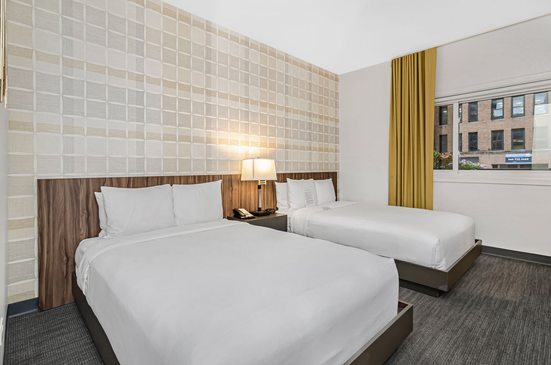 A hotel room with two white-bedded queen beds, beige patterned walls, a dark wood headboard, and mustard-colored curtains.