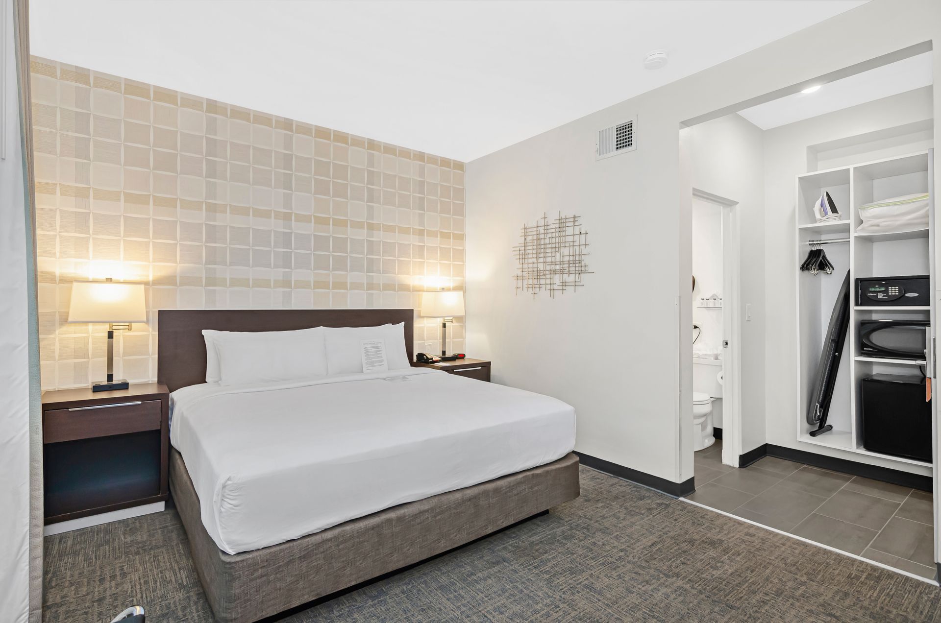 A modern hotel room with a king bed, bedside tables with lamps, a decorative wall piece, and an open closet area.