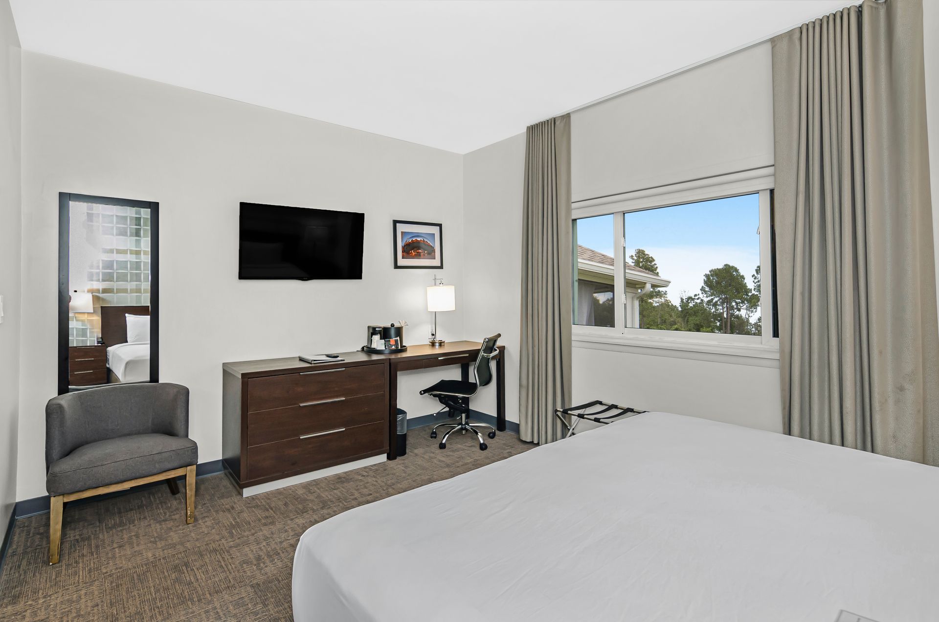 A hotel room with a bed, a desk, a grey armchair, a wall-mounted TV, and a window with floor-length beige curtains.