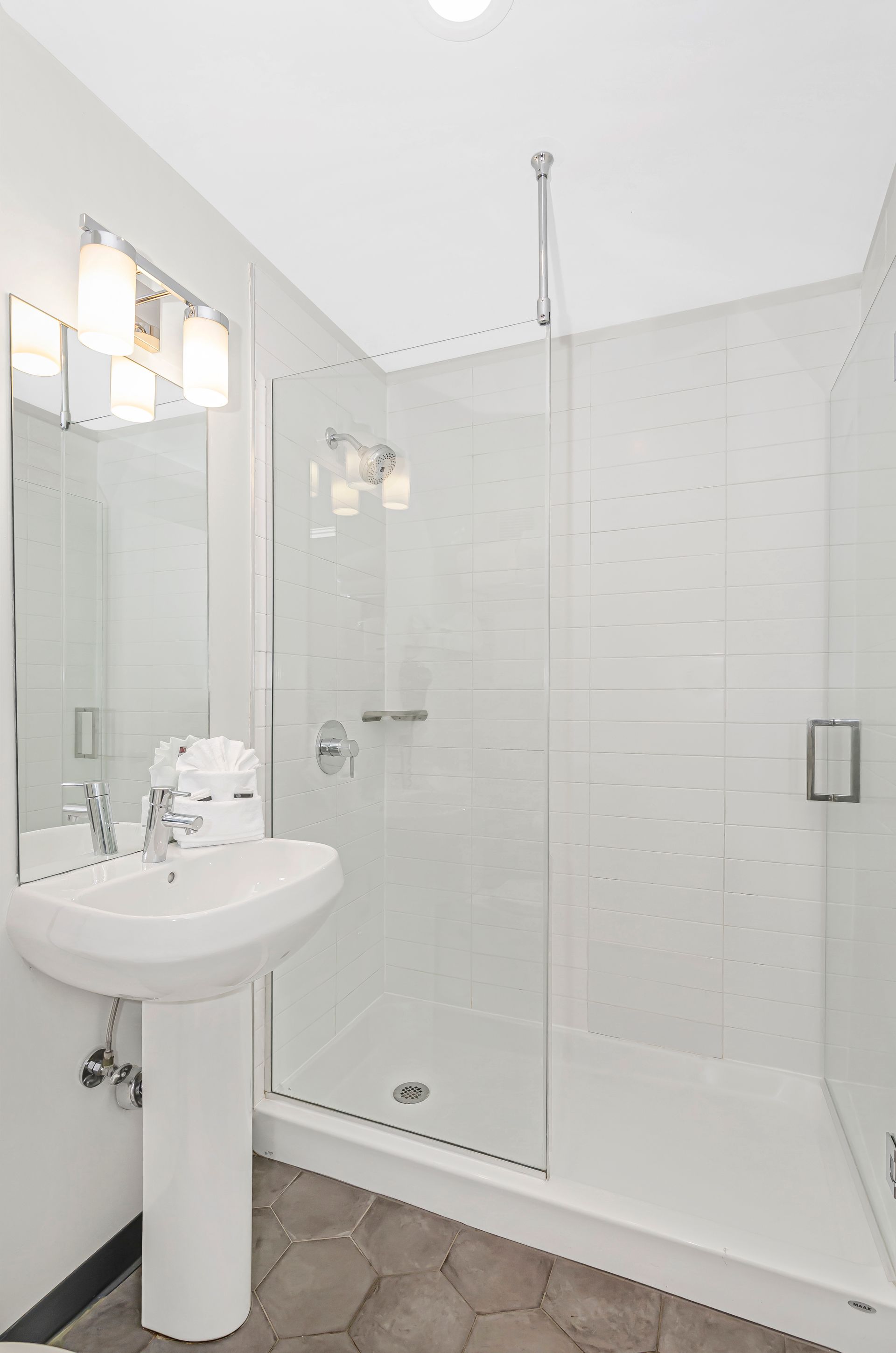 A modern bathroom featuring a white pedestal sink, a glass-enclosed shower, and light gray hexagon floor tiles.
