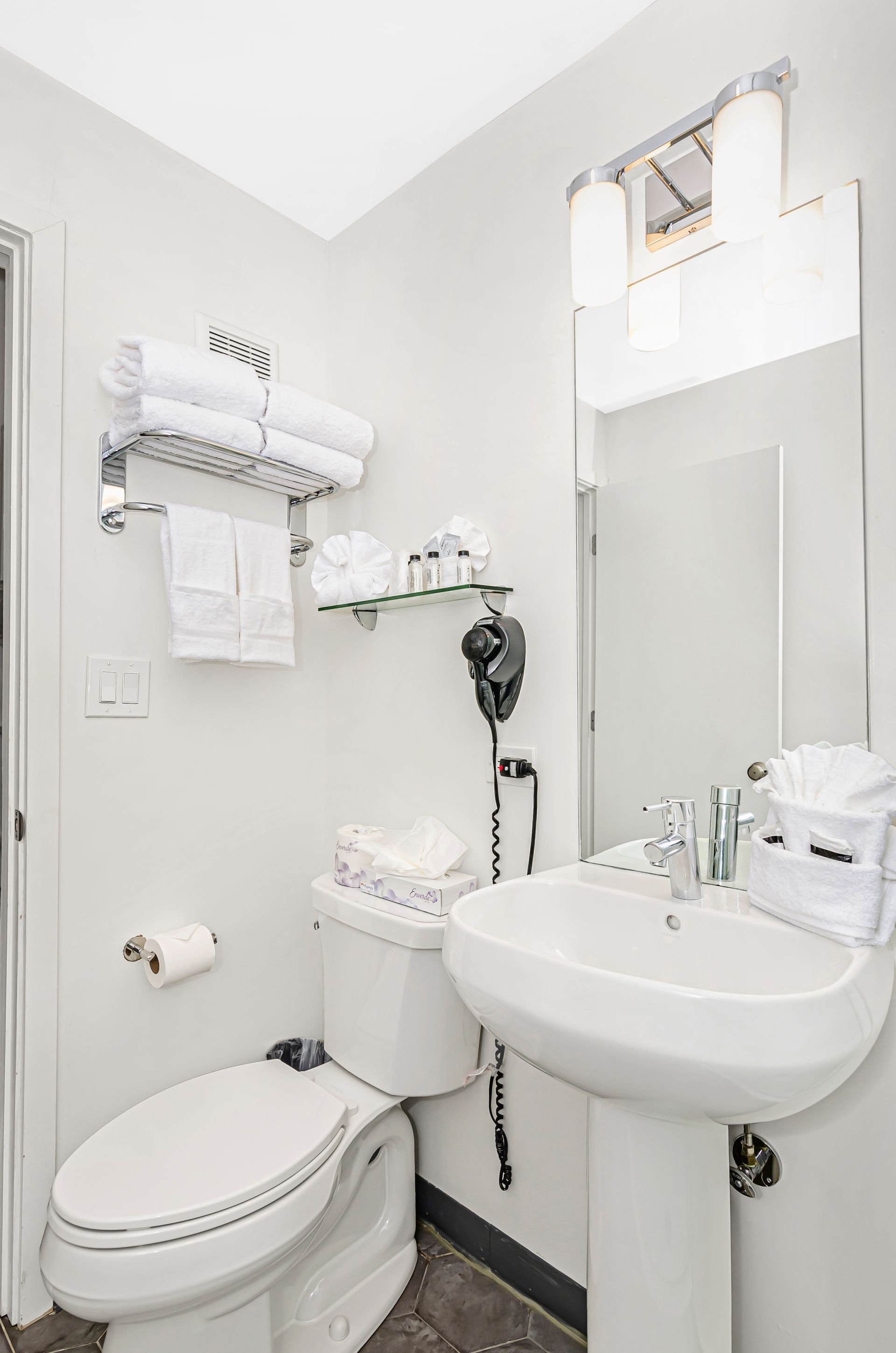 A bright, white bathroom featuring a pedestal sink, toilet, wall-mounted towels, shelves with amenities, and a hairdryer.