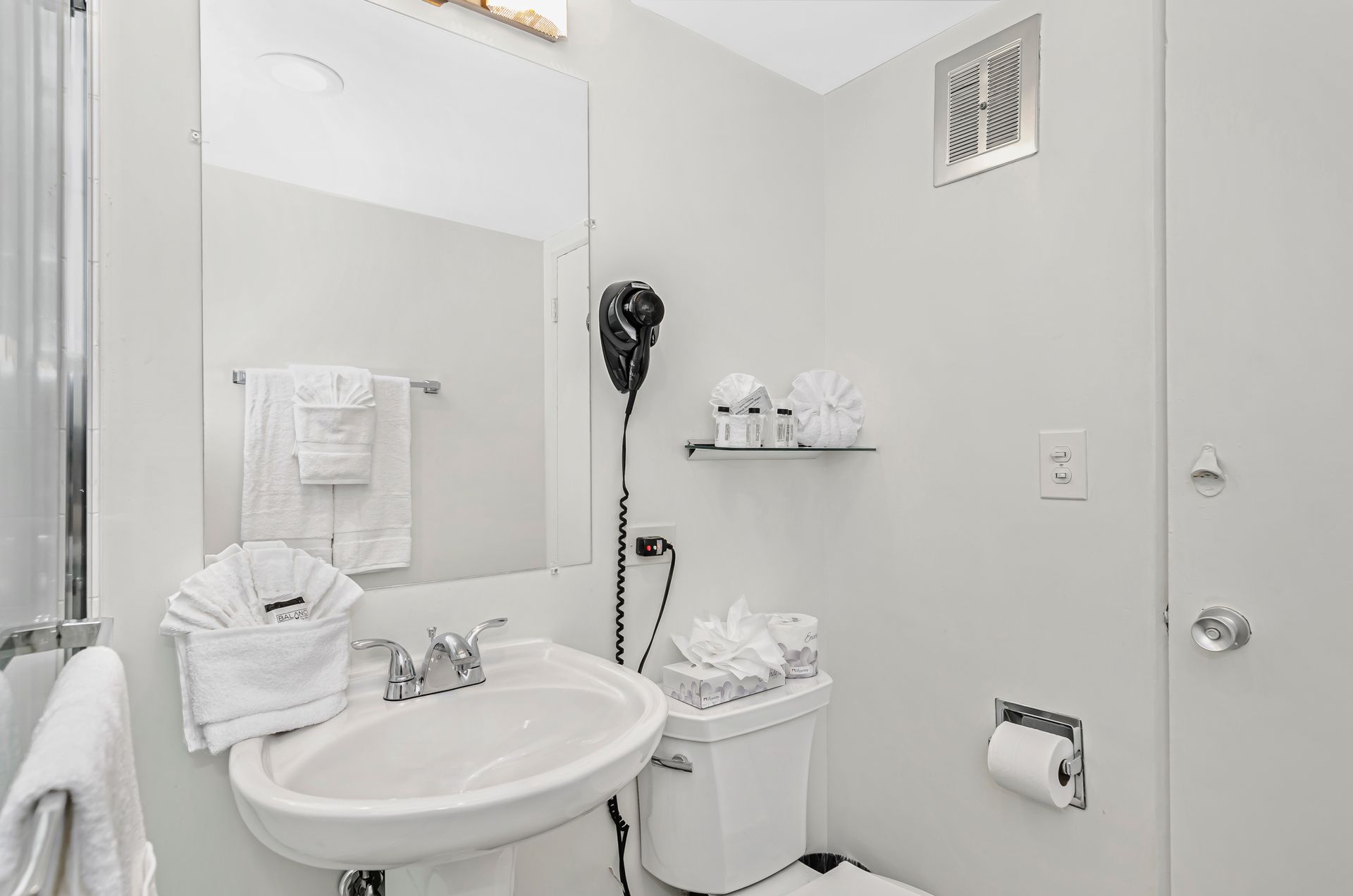 A bright, white bathroom featuring a pedestal sink, a toilet, a wall-mounted hair dryer, and towels on a towel bar.