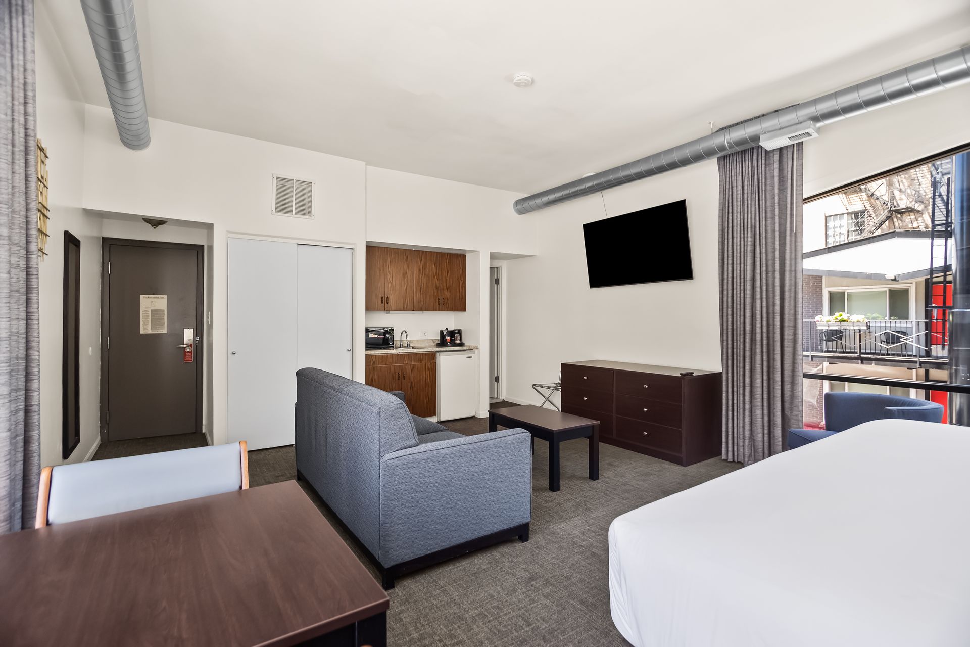 Modern hotel room with two beds, gray sofa, TV, desk, and balcony door.