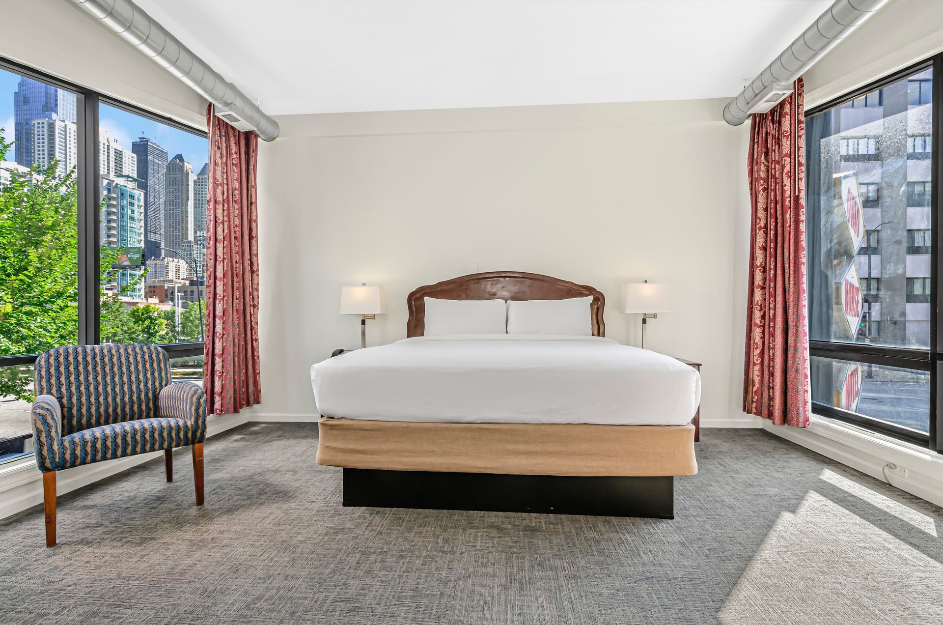 A hotel room with a king bed, grey carpeting, an armchair, and two large windows with city views.