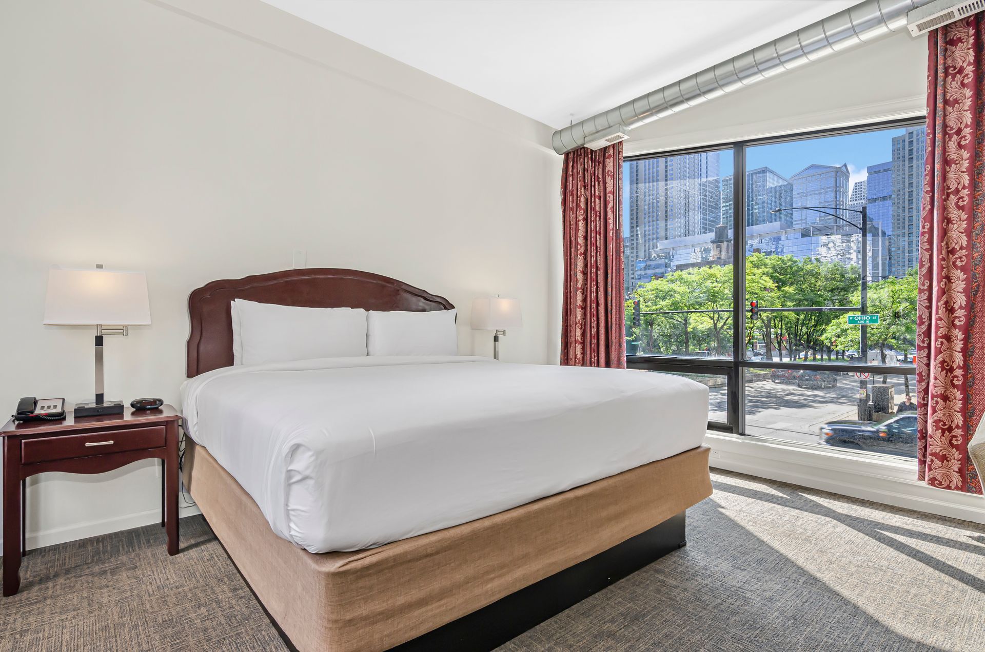 A hotel room featuring a king-size bed with white bedding, a wooden nightstand, and large windows overlooking a city street.