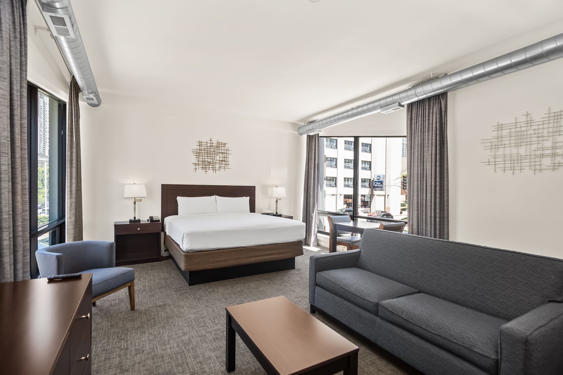 Modern hotel room with a king bed, gray sofa, desk, and balcony doors overlooking city buildings.  Pull Out Sofa 