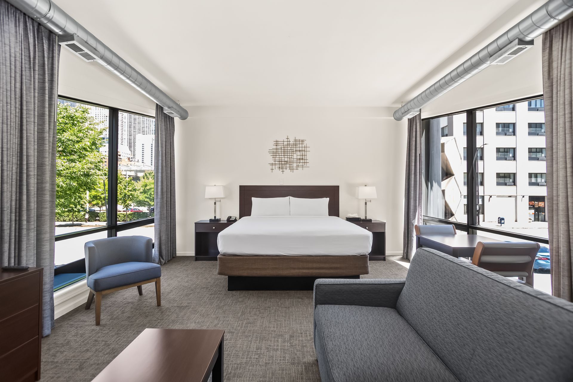 Bright hotel room with a king bed, gray sofa, armchair, and large windows overlooking city buildings