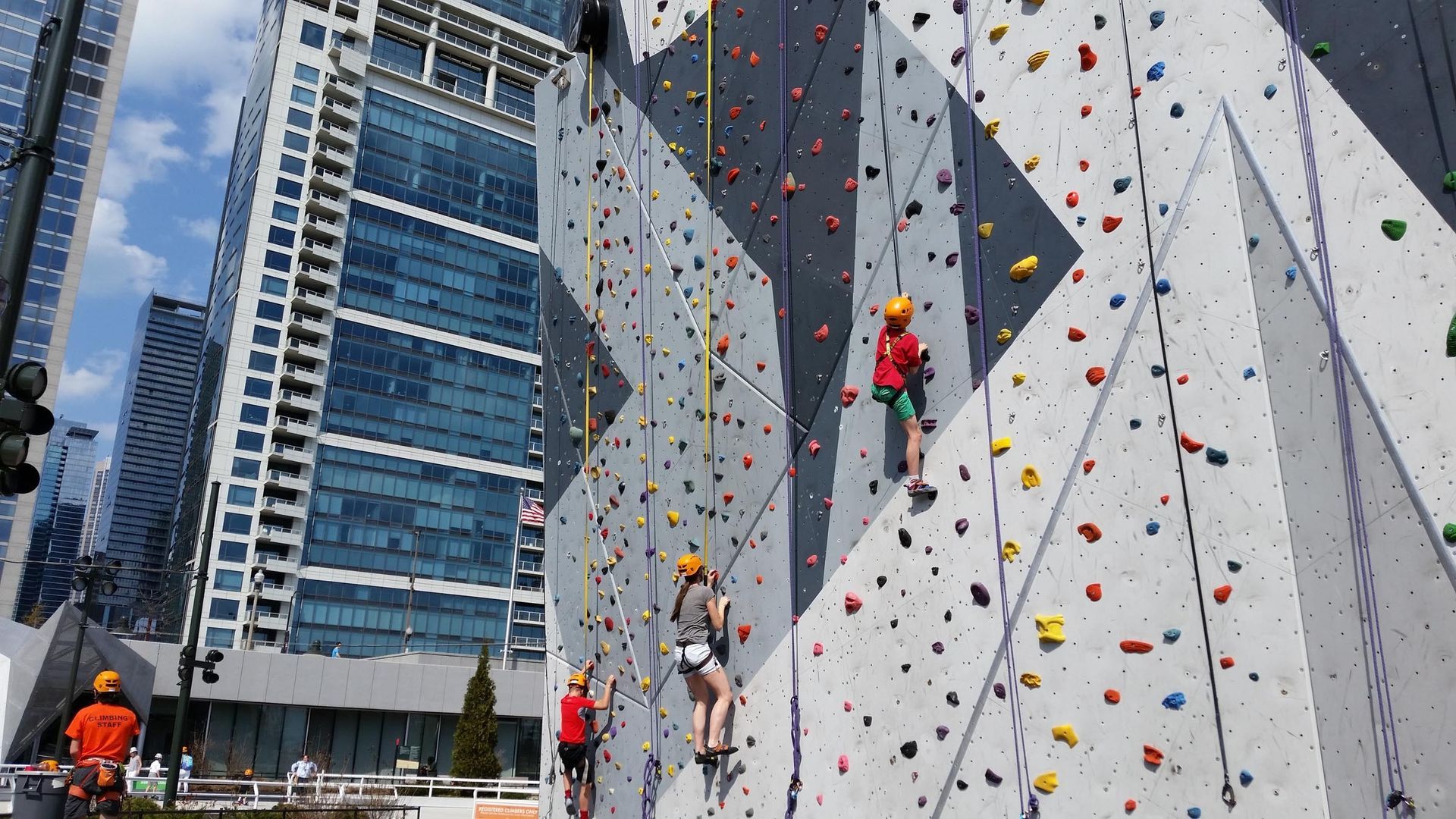 People climbing an outdoor rock wall. Multi-colored handholds, harnesses, and safety ropes. City buildings in background.