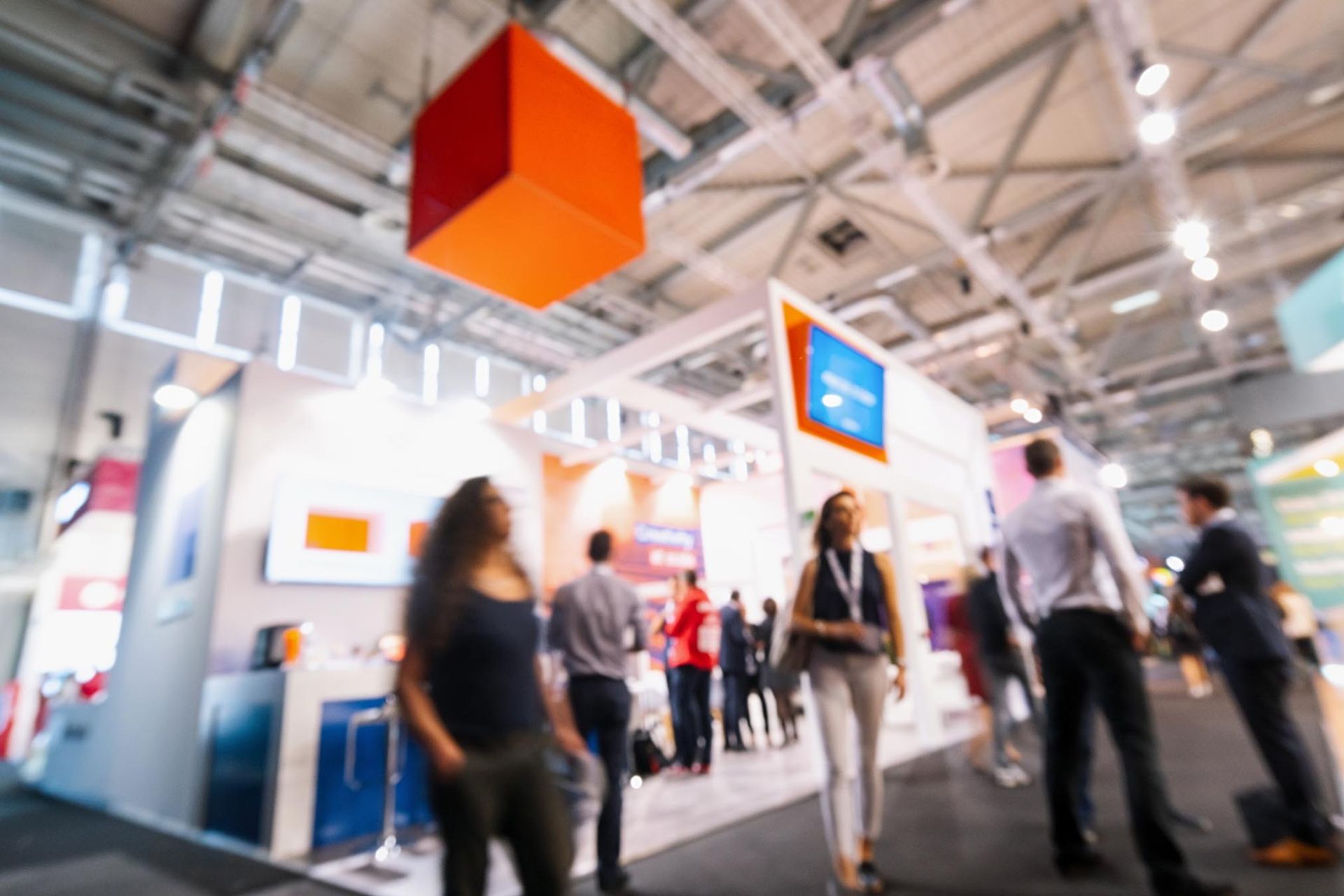 Blurred view of a trade show with people milling about booths; a large orange cube hangs from the ceiling.