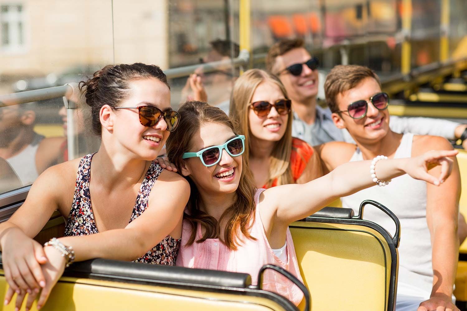 People on a yellow tour bus, pointing, smiling, and wearing sunglasses in a city.