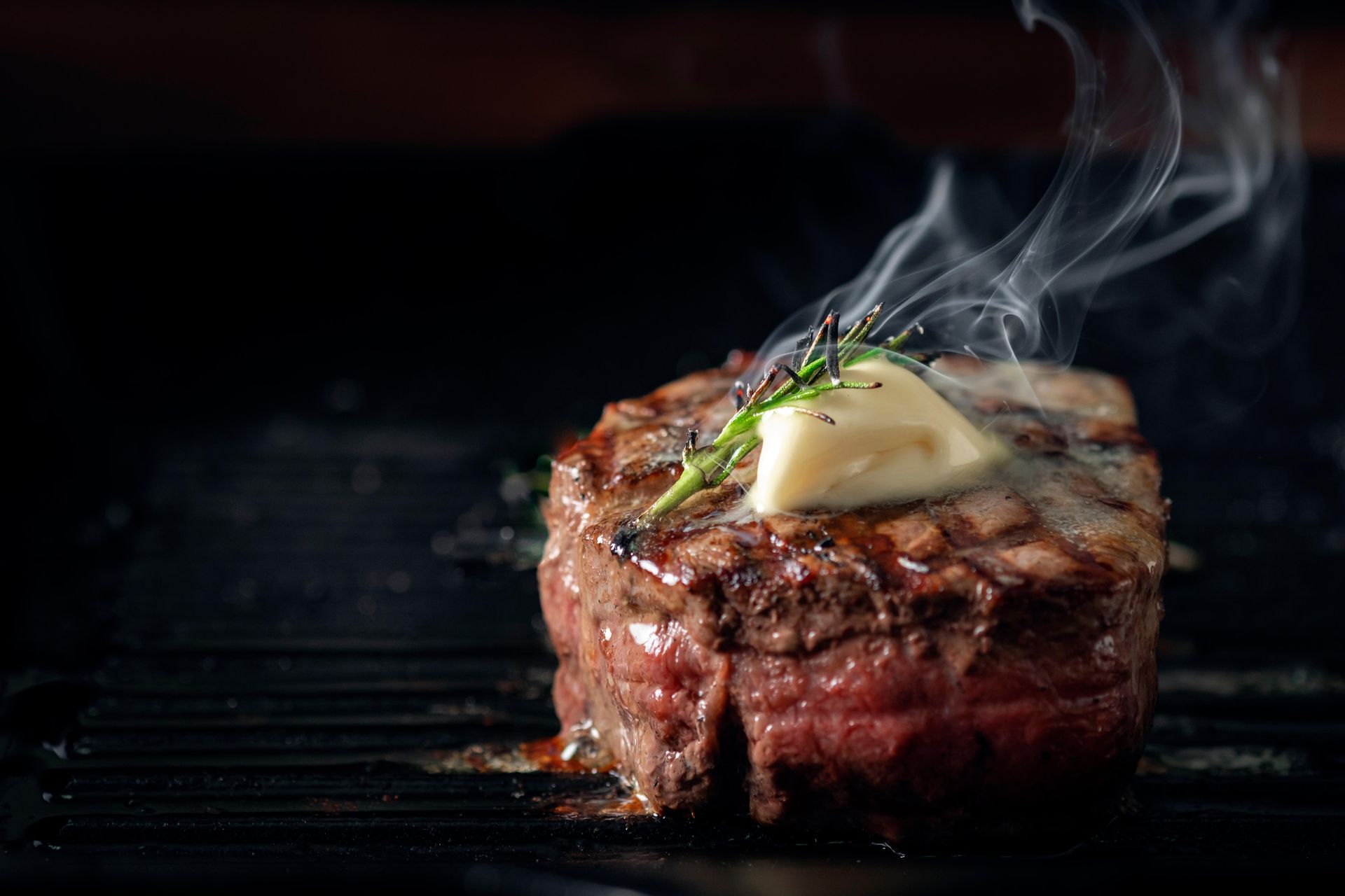 A seared steak topped with a melting pat of butter and fresh herbs, releasing wisps of steam on a black grill pan.