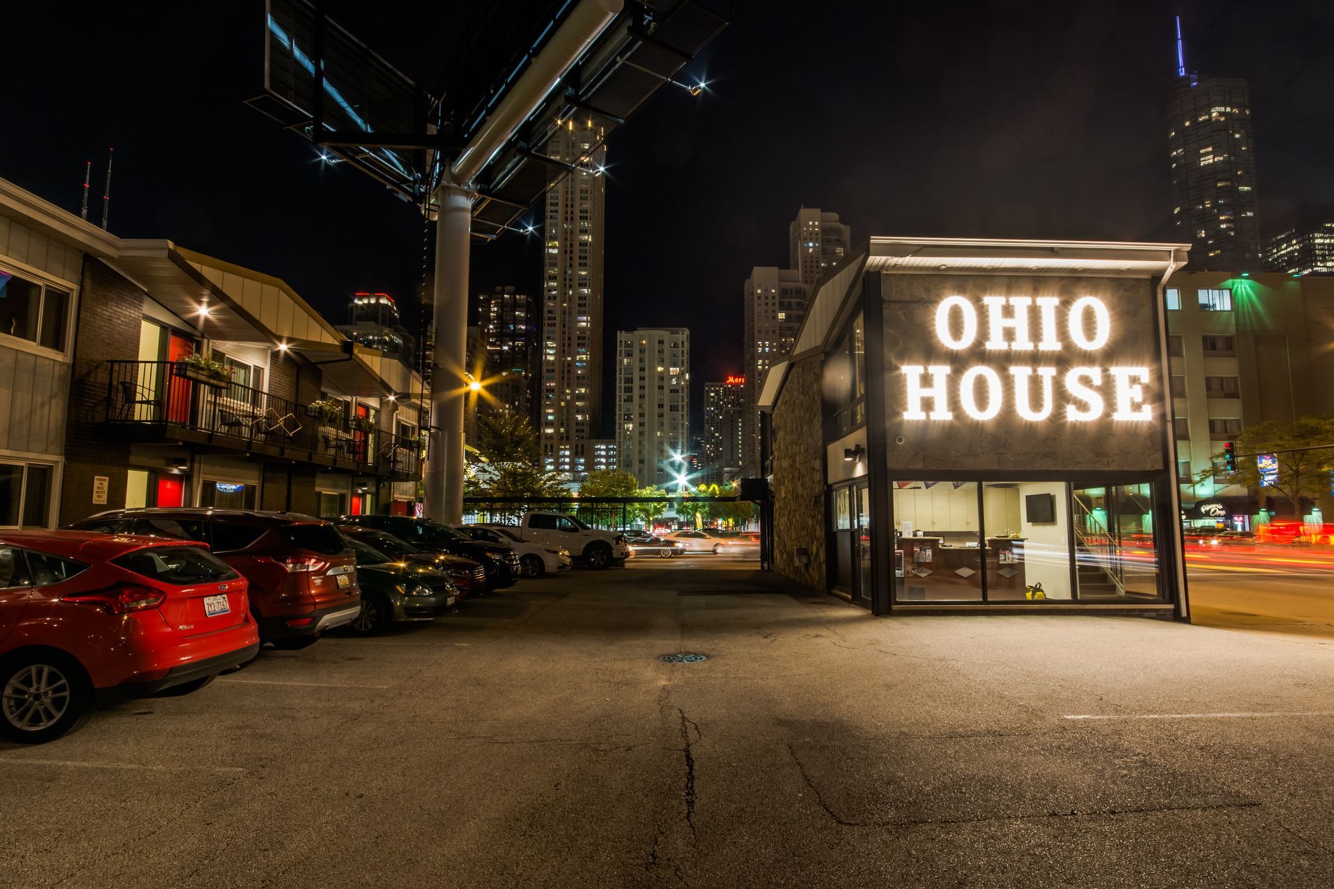 Ohio House bar exterior at night, with illuminated sign, parked cars, and Chicago skyline.
