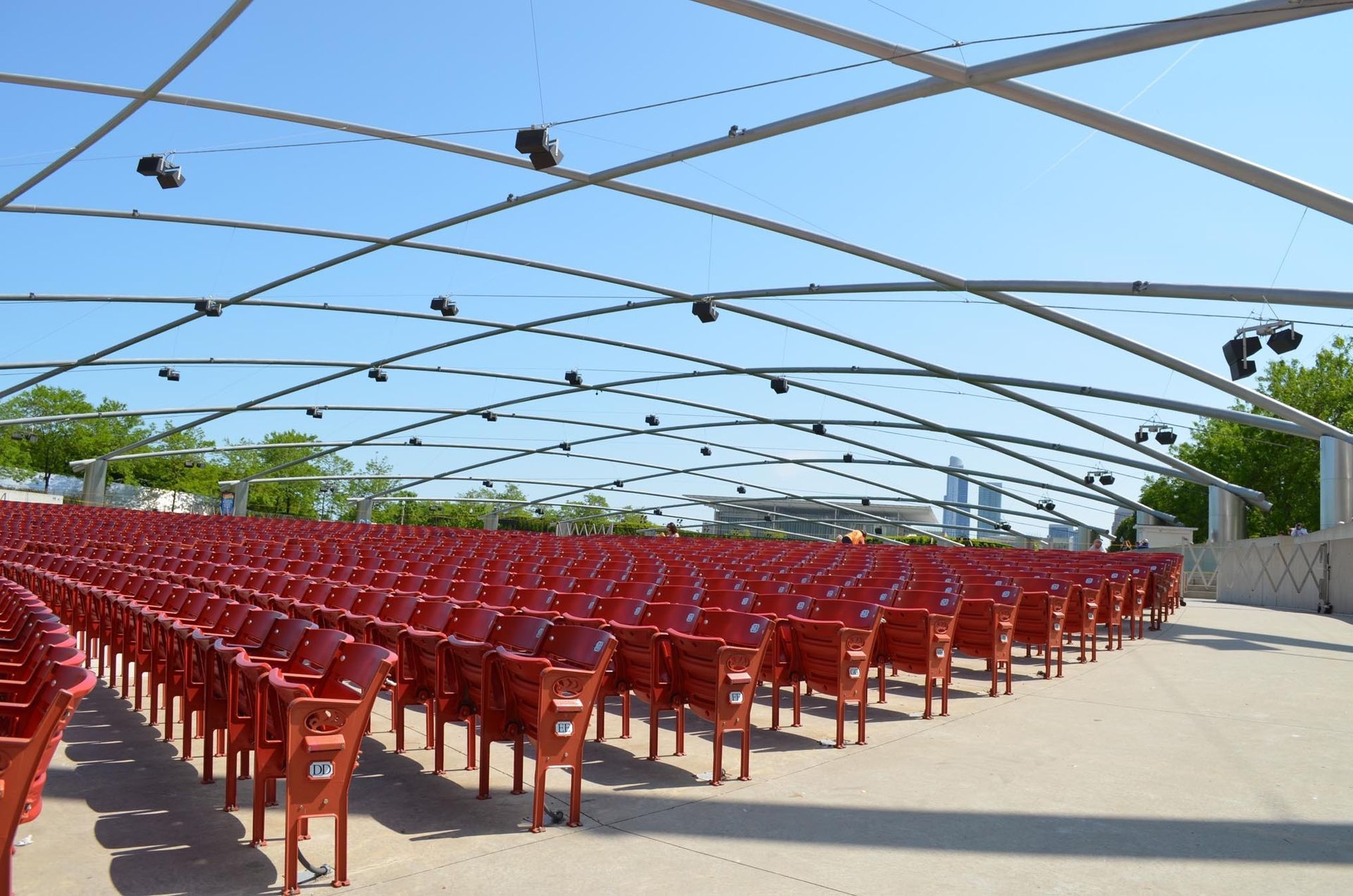 Rows of empty red seats under a curved metal canopy at an outdoor amphitheater on a sunny day.