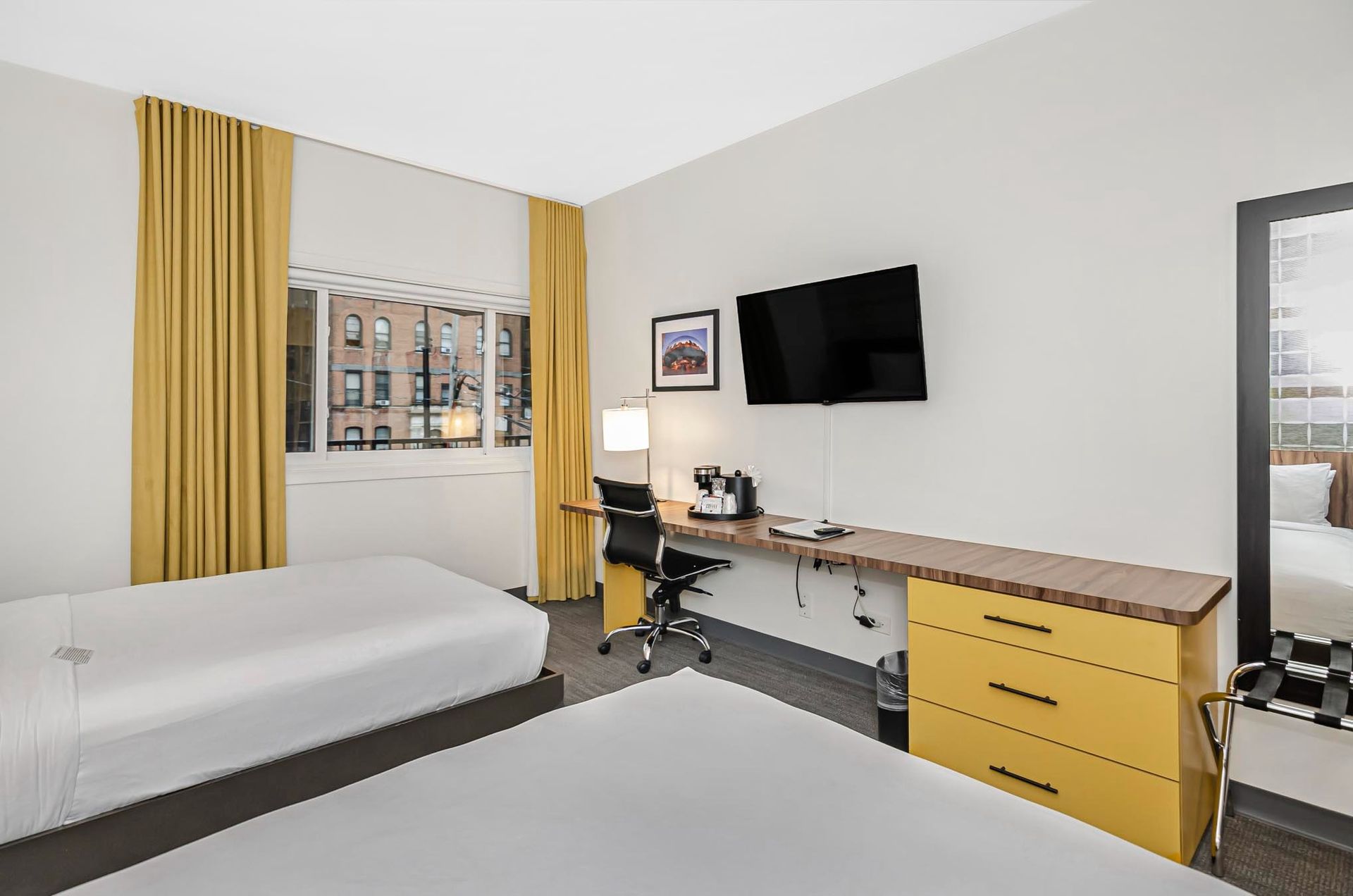 Hotel room with two beds, desk with TV, large mirror, and window with yellow curtains.