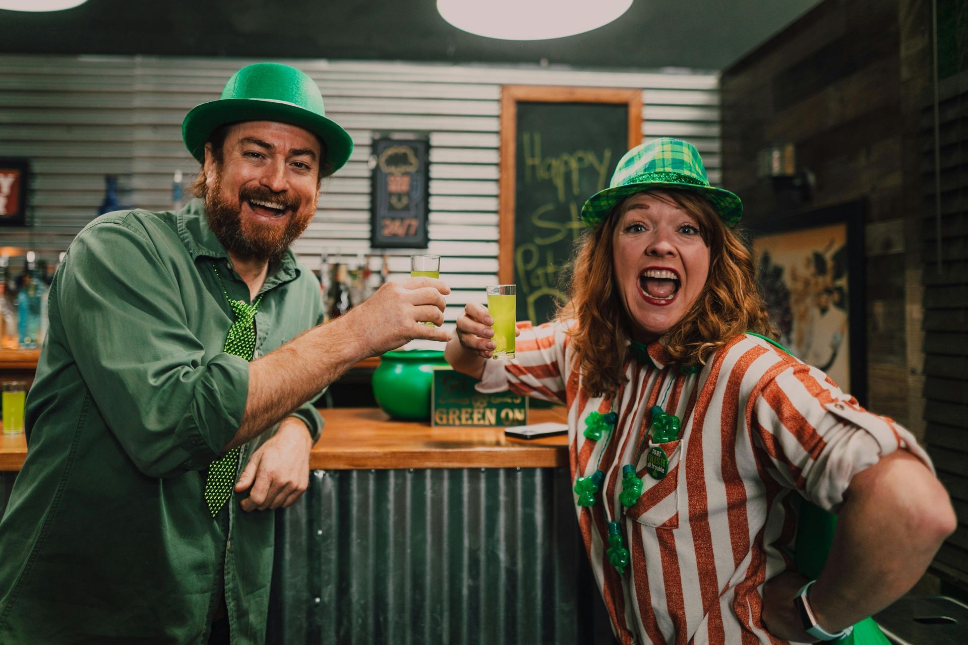 Two people in green hats toast with small glasses in a festive bar setting.