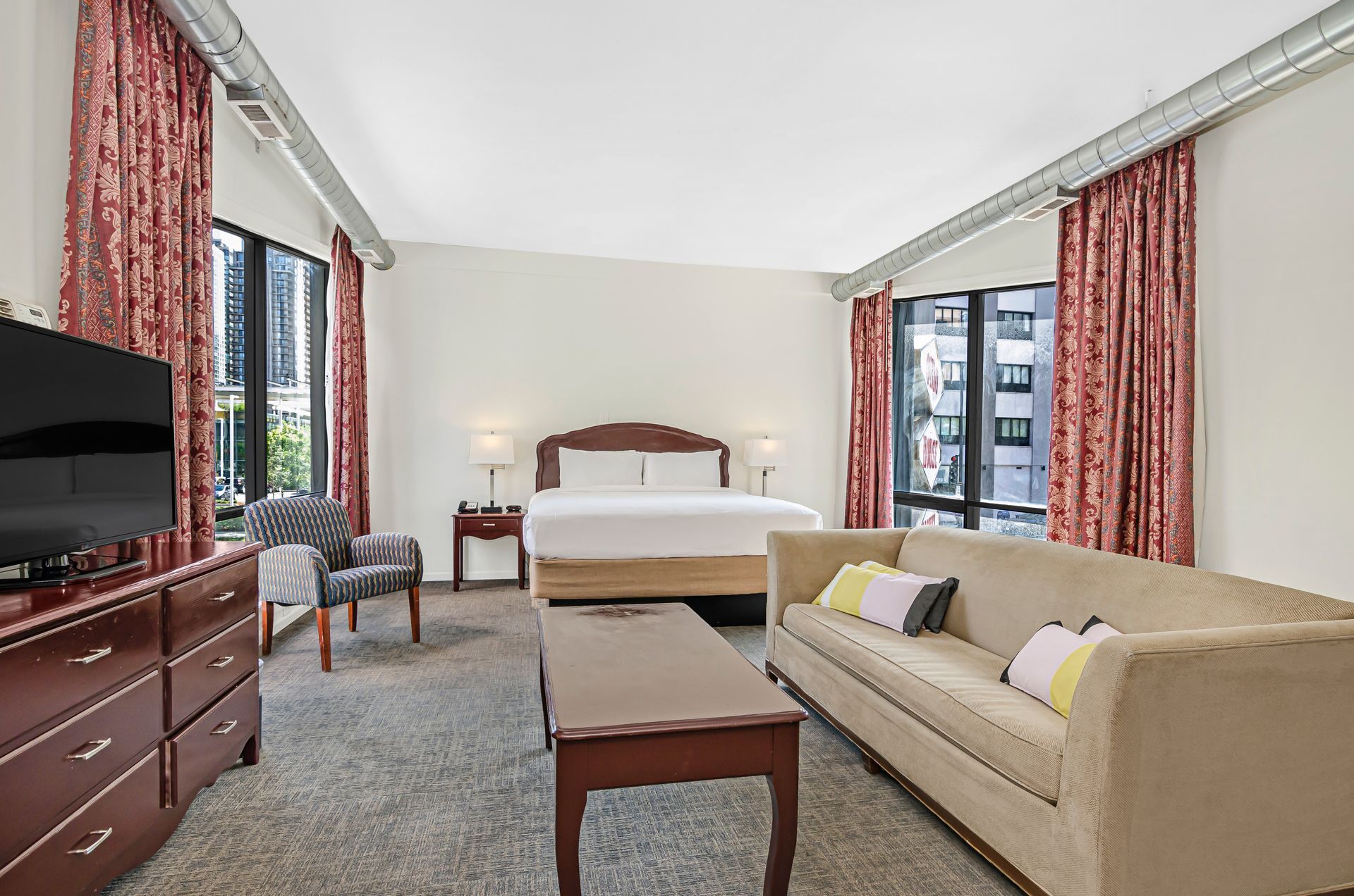 A hotel room with a queen bed, a beige sofa, a coffee table, a grey armchair, a dresser with a TV, and red curtains.