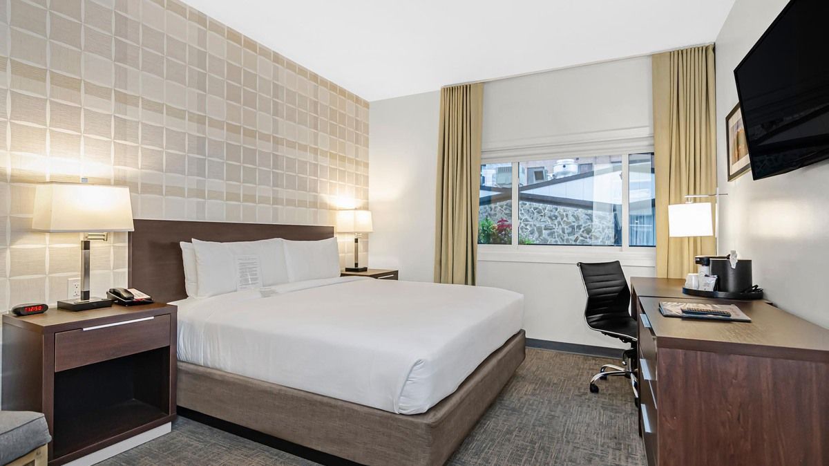 Hotel room with bed, nightstand, desk, and window. Neutral colors.