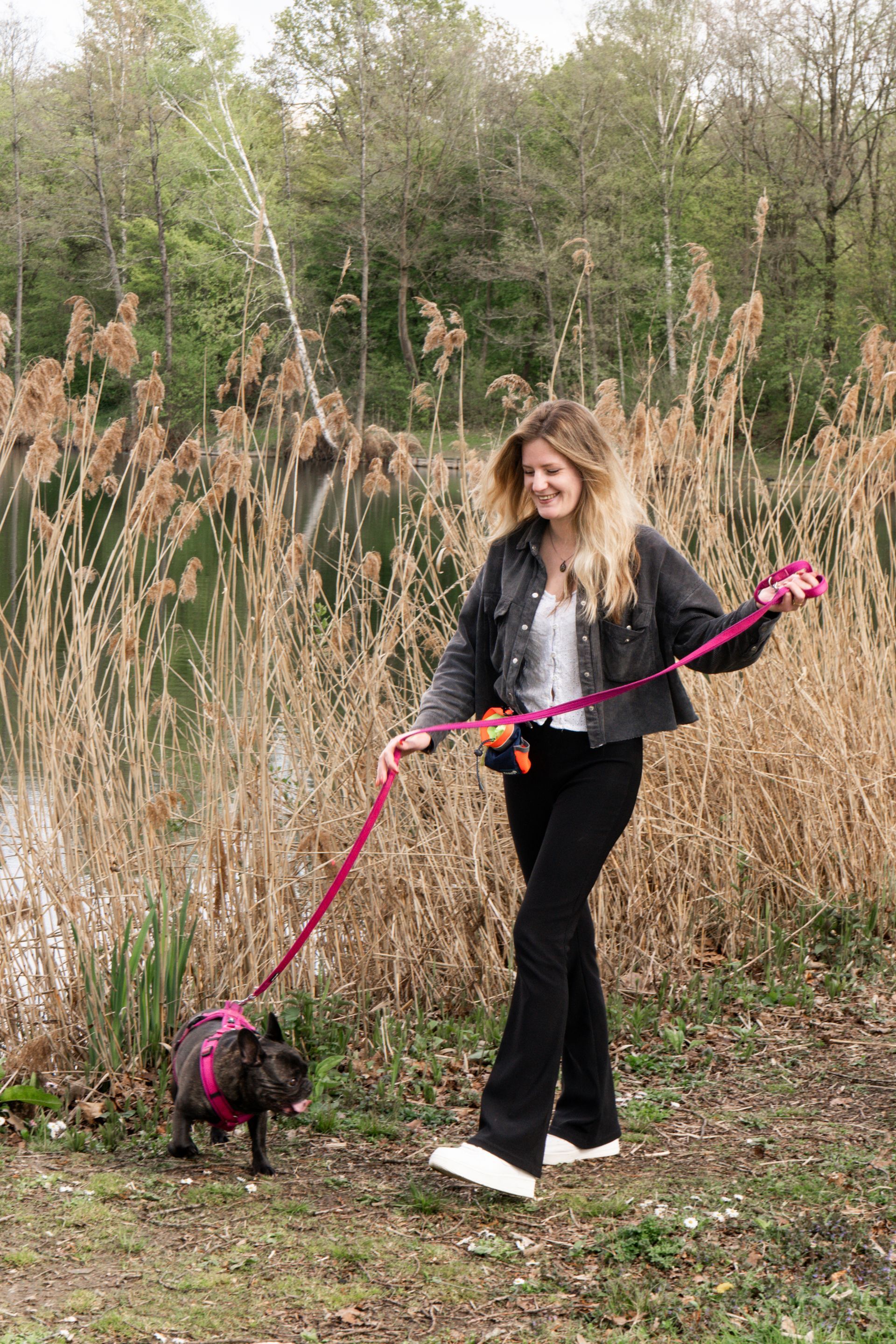 Woman walking a small dog on a pink leash in a grassy outdoor field.