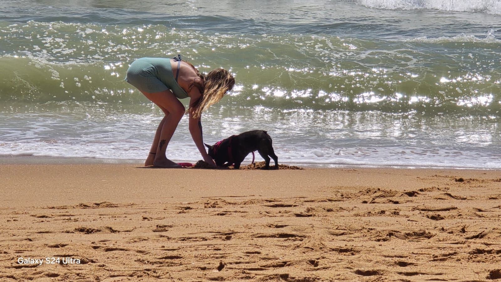 Woman bends toward a black dog on a sandy beach by the ocean.