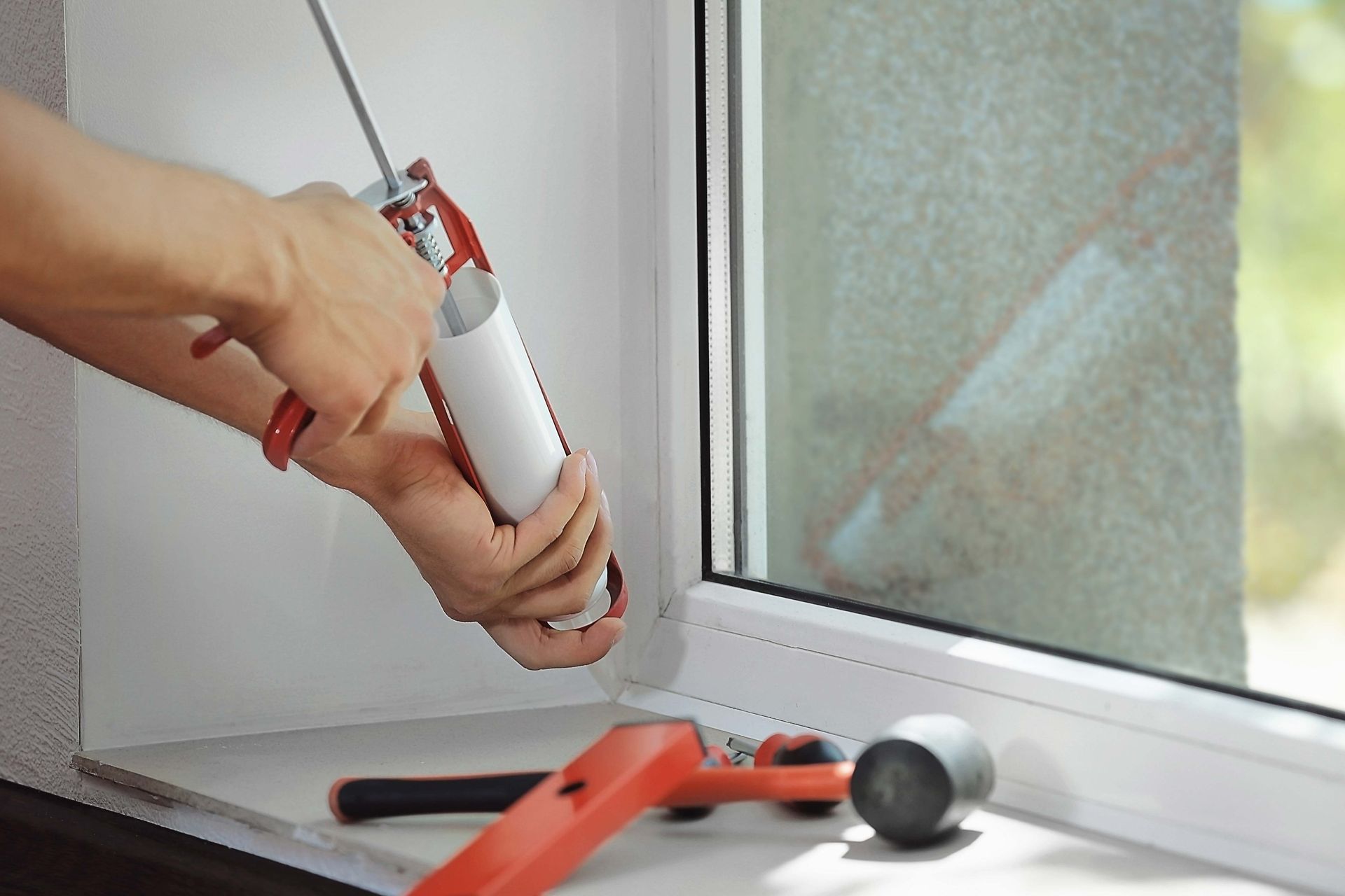 Person caulking a window frame with a caulk gun. White frame and caulk gun against the window and sill.