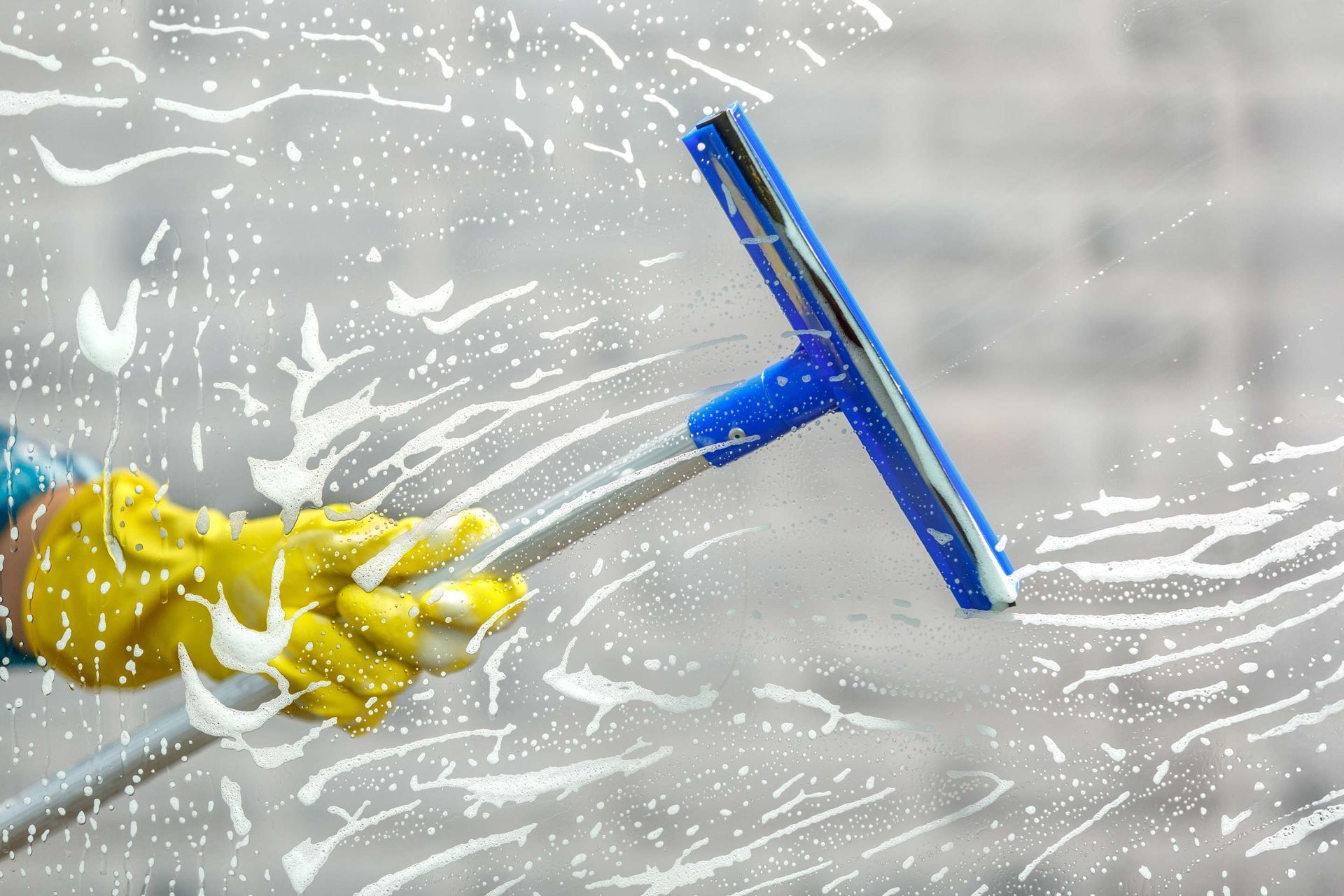 A hand in a yellow glove uses a blue squeegee on a soapy window.
