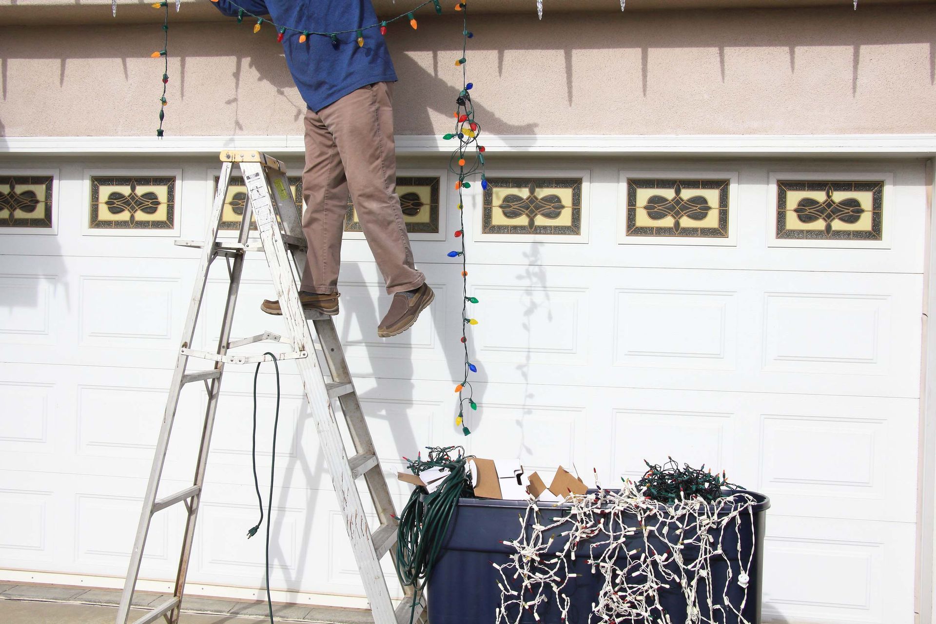 Person on ladder hangs Christmas lights on a white garage.