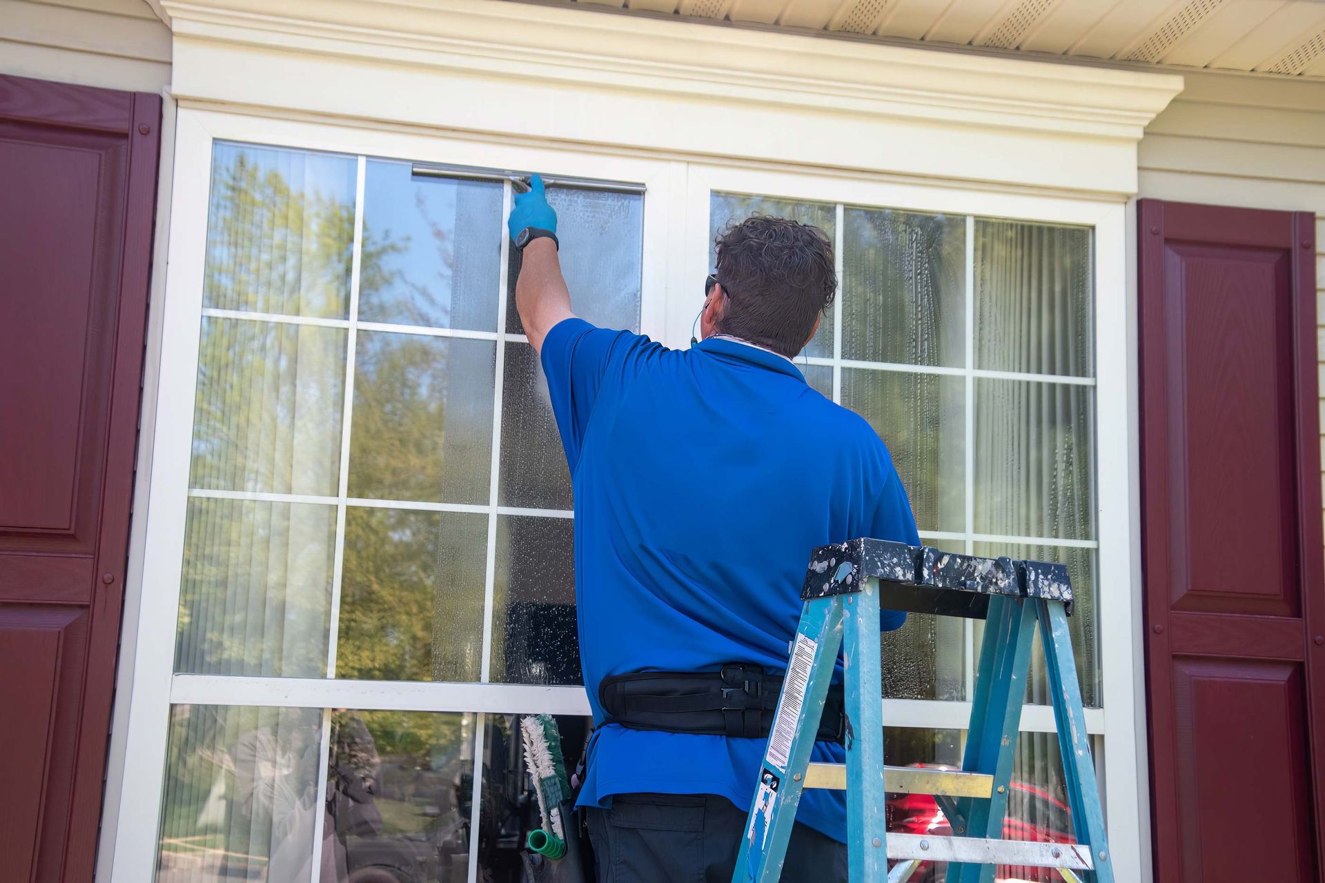 Person cleaning a window with a squeegee on a ladder; white window frame, blue shirt, red shutters.