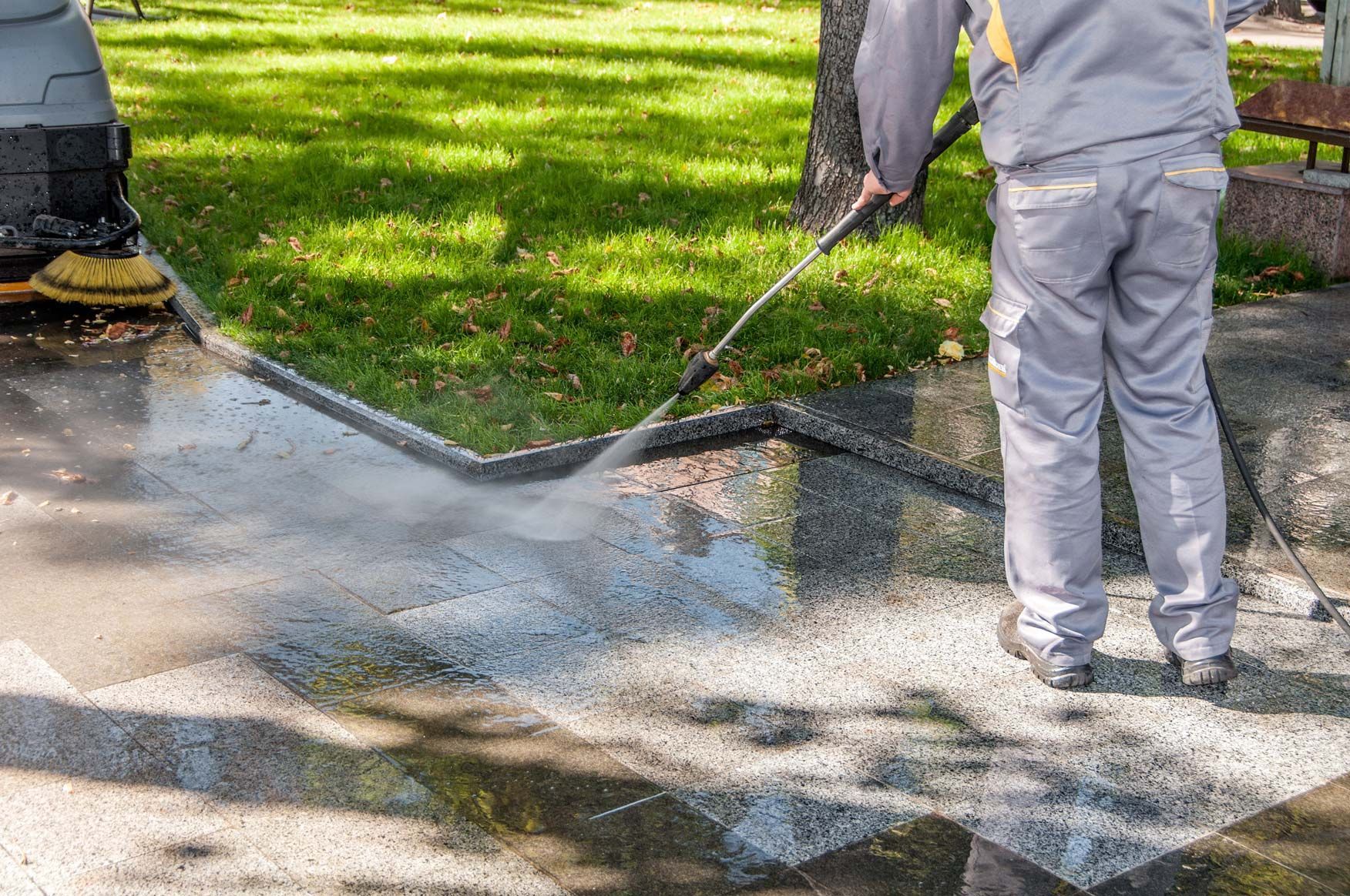 Worker pressure washing a concrete sidewalk next to a grassy area, creating water spray.