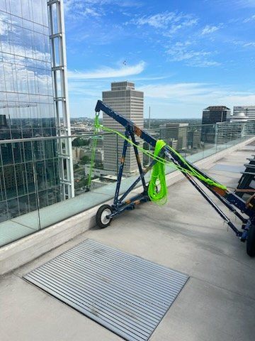 Crane on a rooftop with a cityscape backdrop. Green rope hangs from the crane. Blue sky overhead.