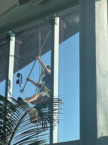 Person suspended on ropes cleaning a large exterior window. Bright blue sky is visible.