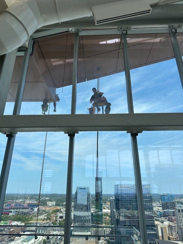 Window washer working on a tall building, seen through other windows; blue sky.