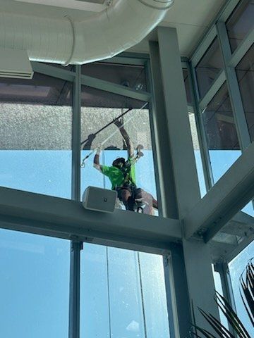Person suspended from a cable, cleaning the exterior of a large window. Green shirt, harness, and skyscraper setting.