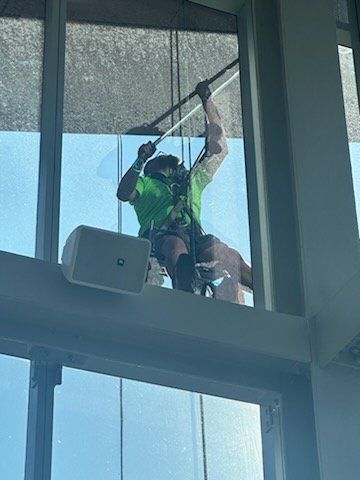 Window washer suspended, cleaning a high-rise window with a long-handled tool. He is wearing a harness and green shirt.