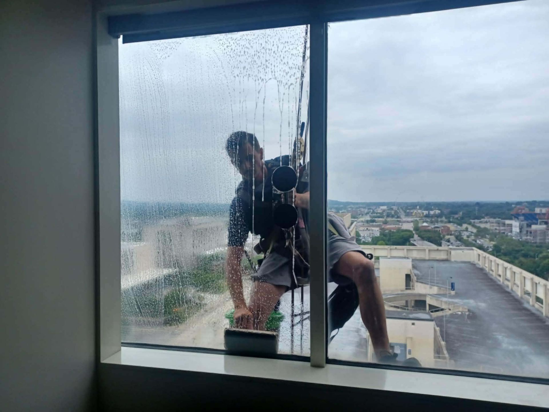Window washer hanging on ropes outside high-rise building, squeegeeing a window. City skyline in the background.