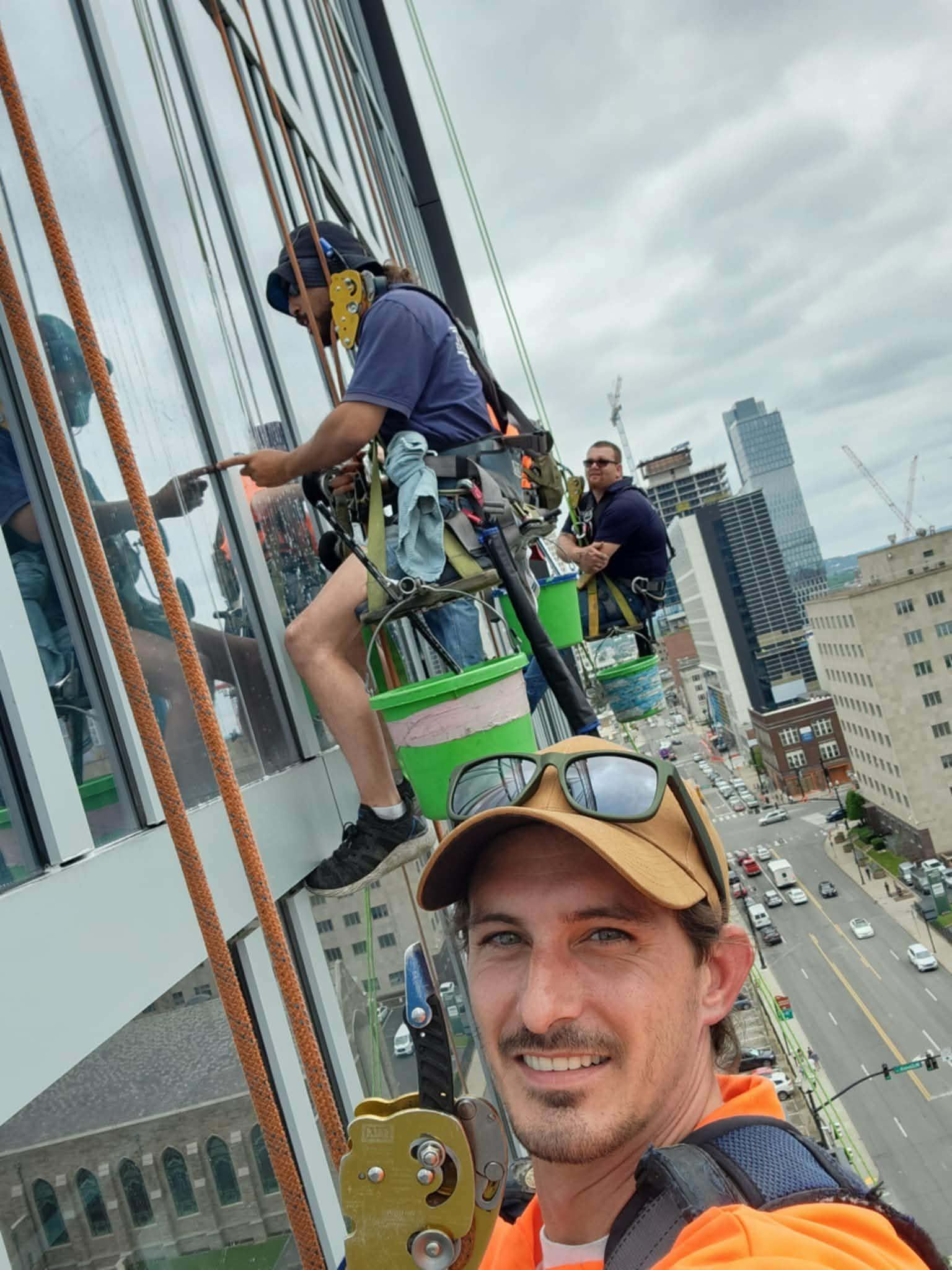 Window washers on a high-rise building with safety gear. Man taking a selfie with cityscape background.