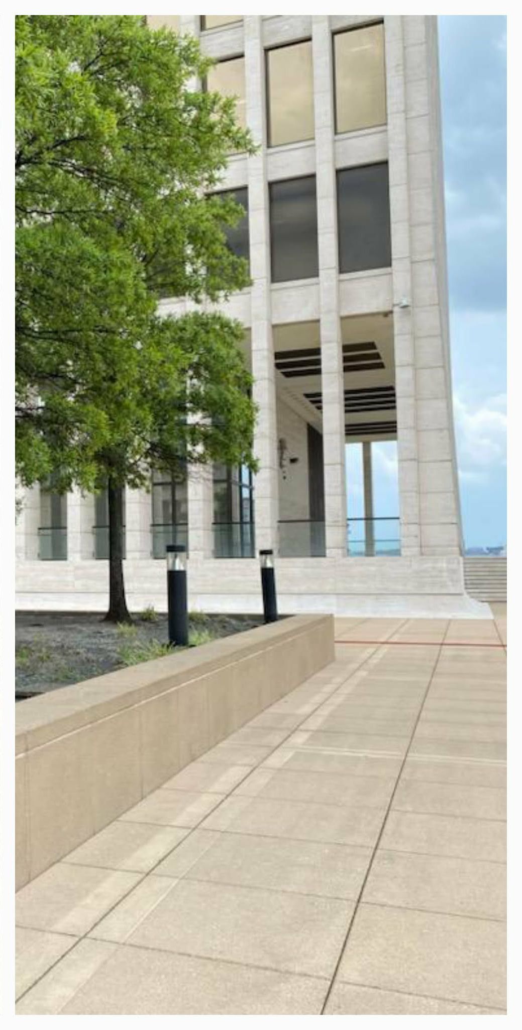 A light-colored building with large windows and a green tree in front. A stone walkway is in the foreground.