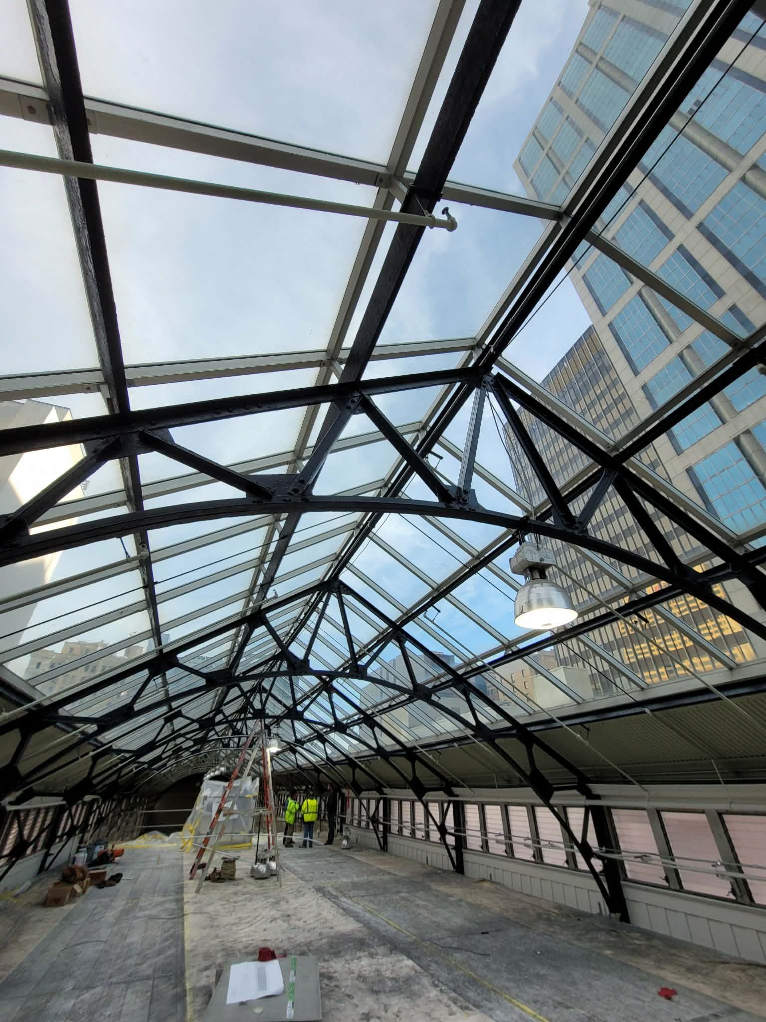 Glass roof structure with exposed beams, construction in progress, city buildings in the background.