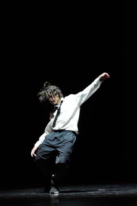 Ballerino con camicia bianca e pantaloni scuri in posa dinamica su un palco buio sotto un riflettore