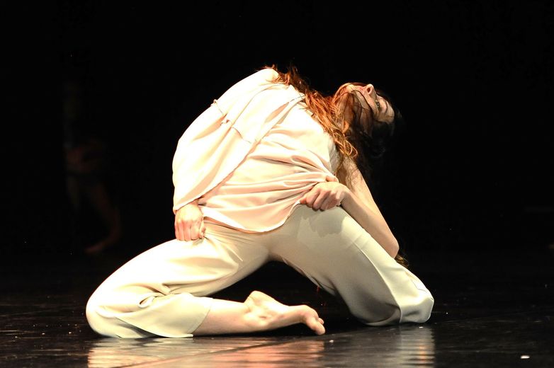 Una ballerina vestita di bianco si accovaccia su un palcoscenico buio, sporgendosi in avanti sotto un caldo riflettore.