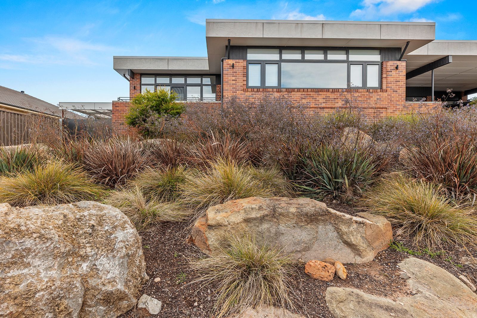 Modern brick house with carport; landscaping in front with rocks and various plants.