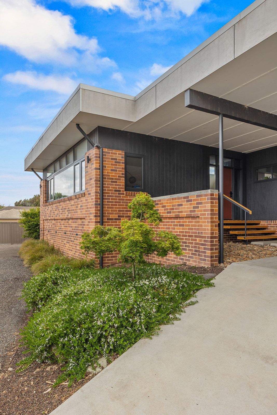 Modern home exterior with brick and dark siding, concrete path, and landscaping under a blue sky.