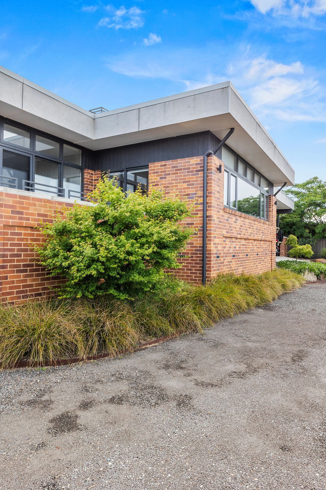 Brick building with large windows and a flat roof, gravel path, and green foliage against a blue sky.