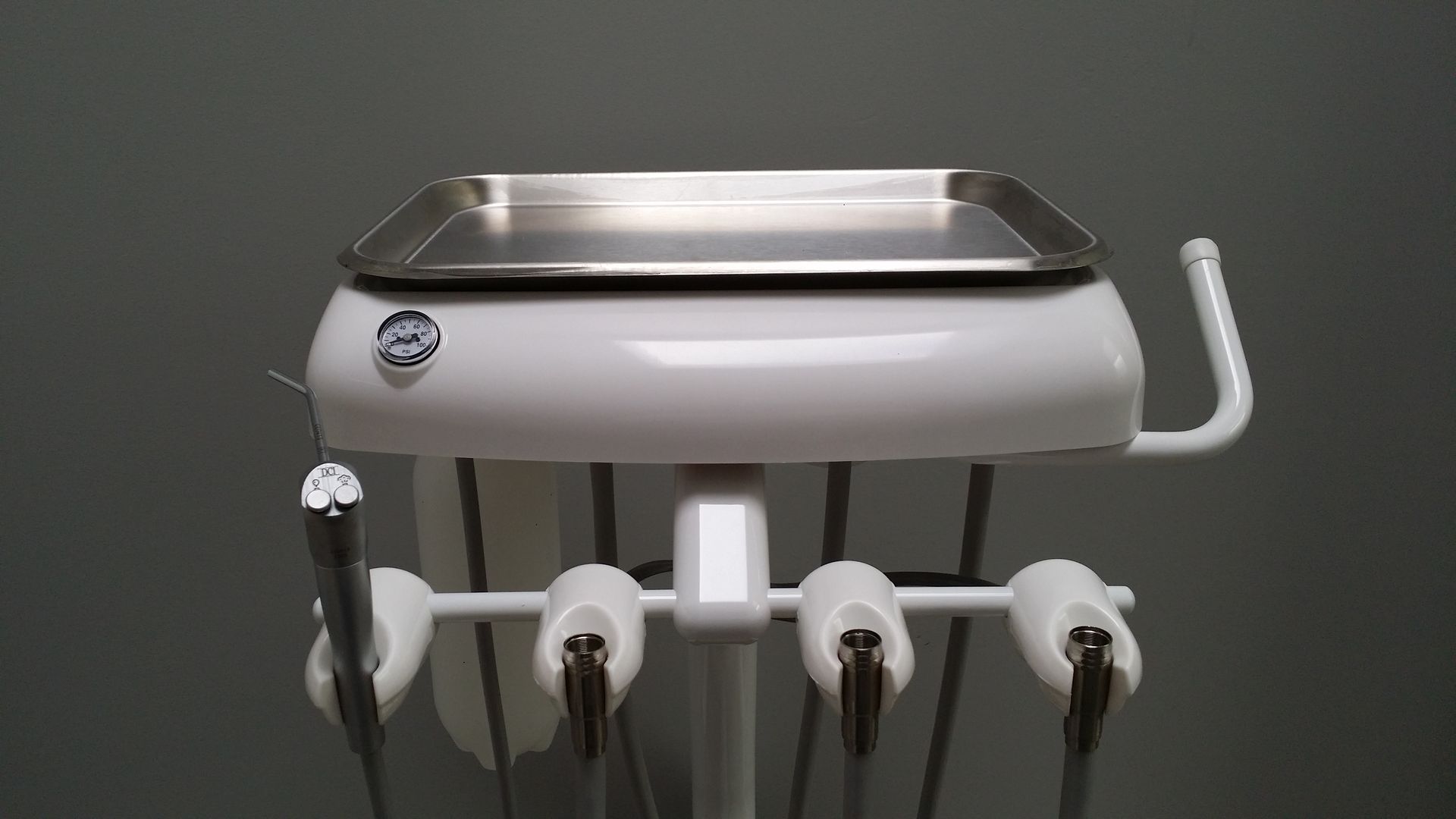 White dental equipment with tray, tools, and hoses in front of a gray wall.