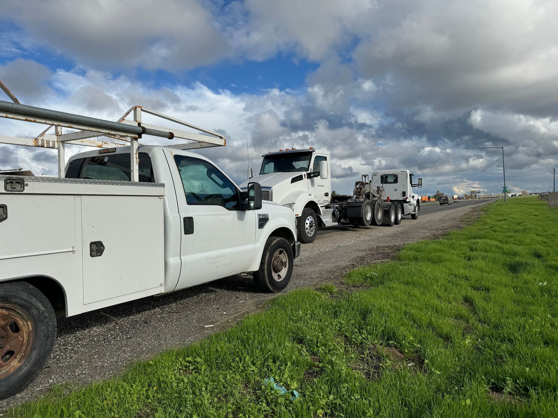 White pickup truck and semi-truck parked on a dirt road beside green grass and under a cloudy blue sky.